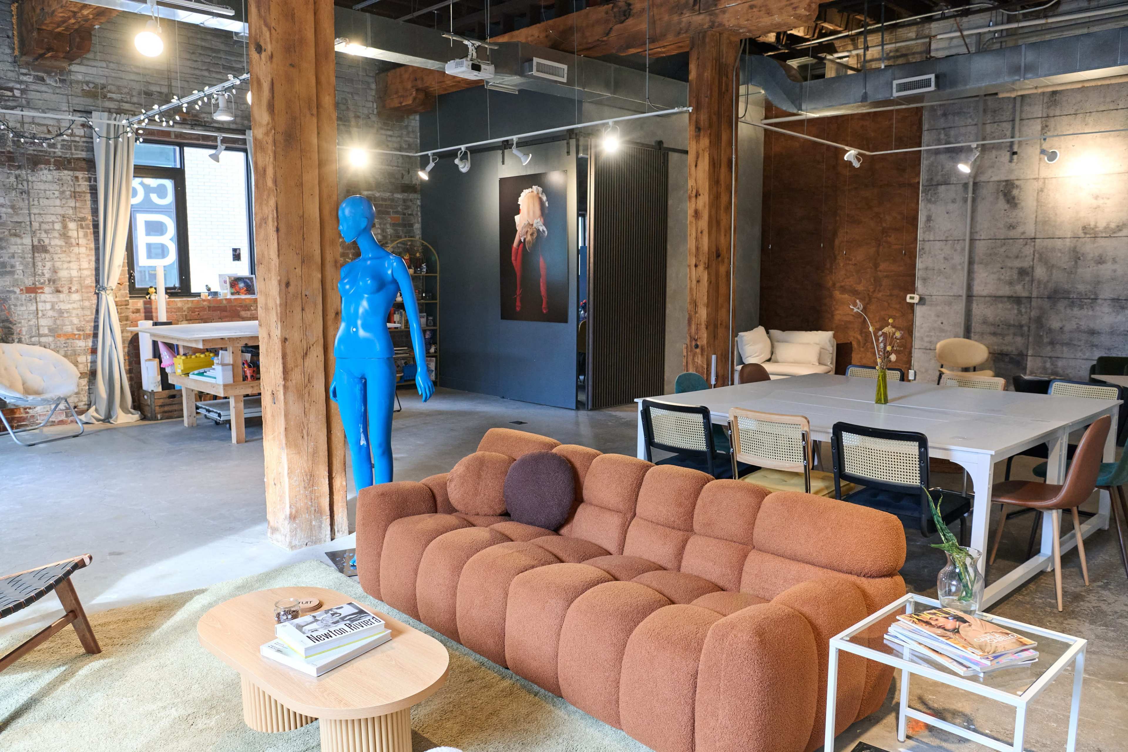 The image shows a modern interior space with a brown sofa, a blue mannequin, and a variety of furniture arranged in an industrial-style setting with exposed beams and concrete walls.