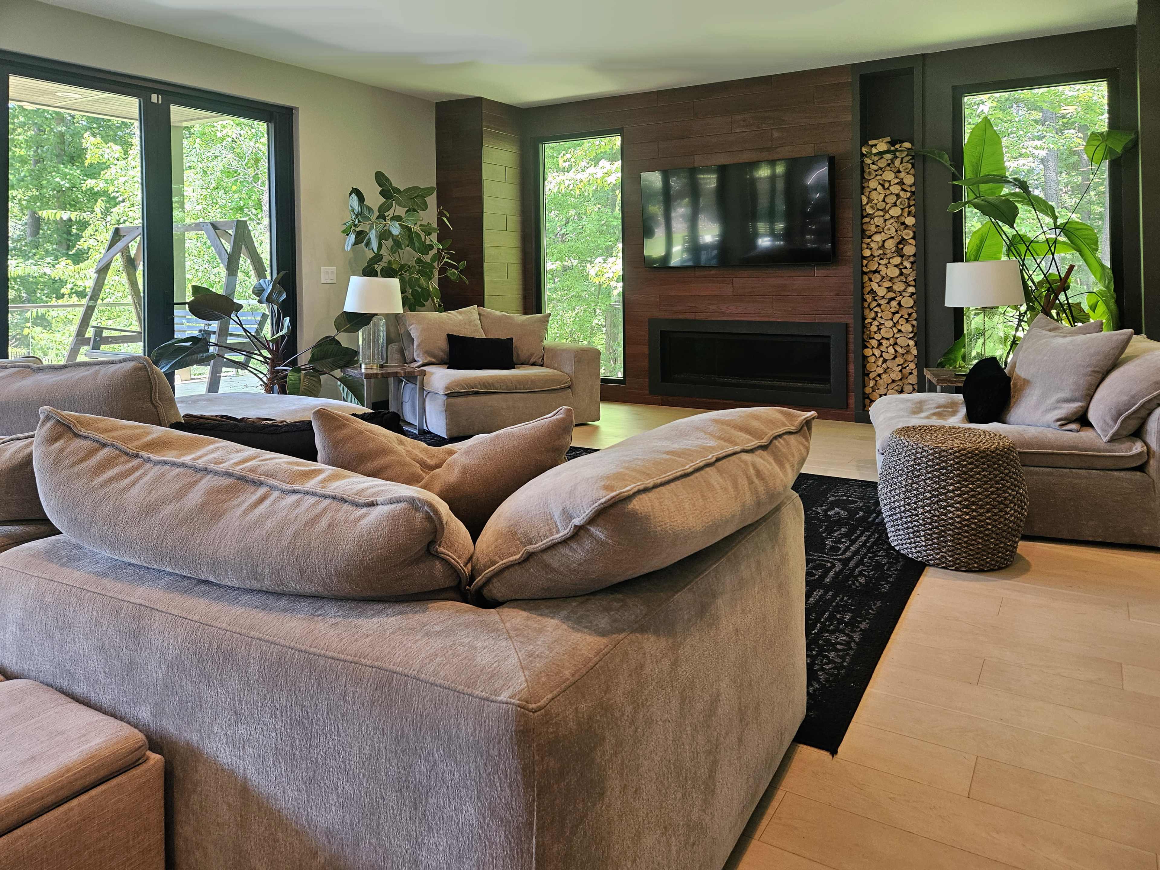 The image shows a modern living room with large windows, cozy seating arrangements, and indoor plants.