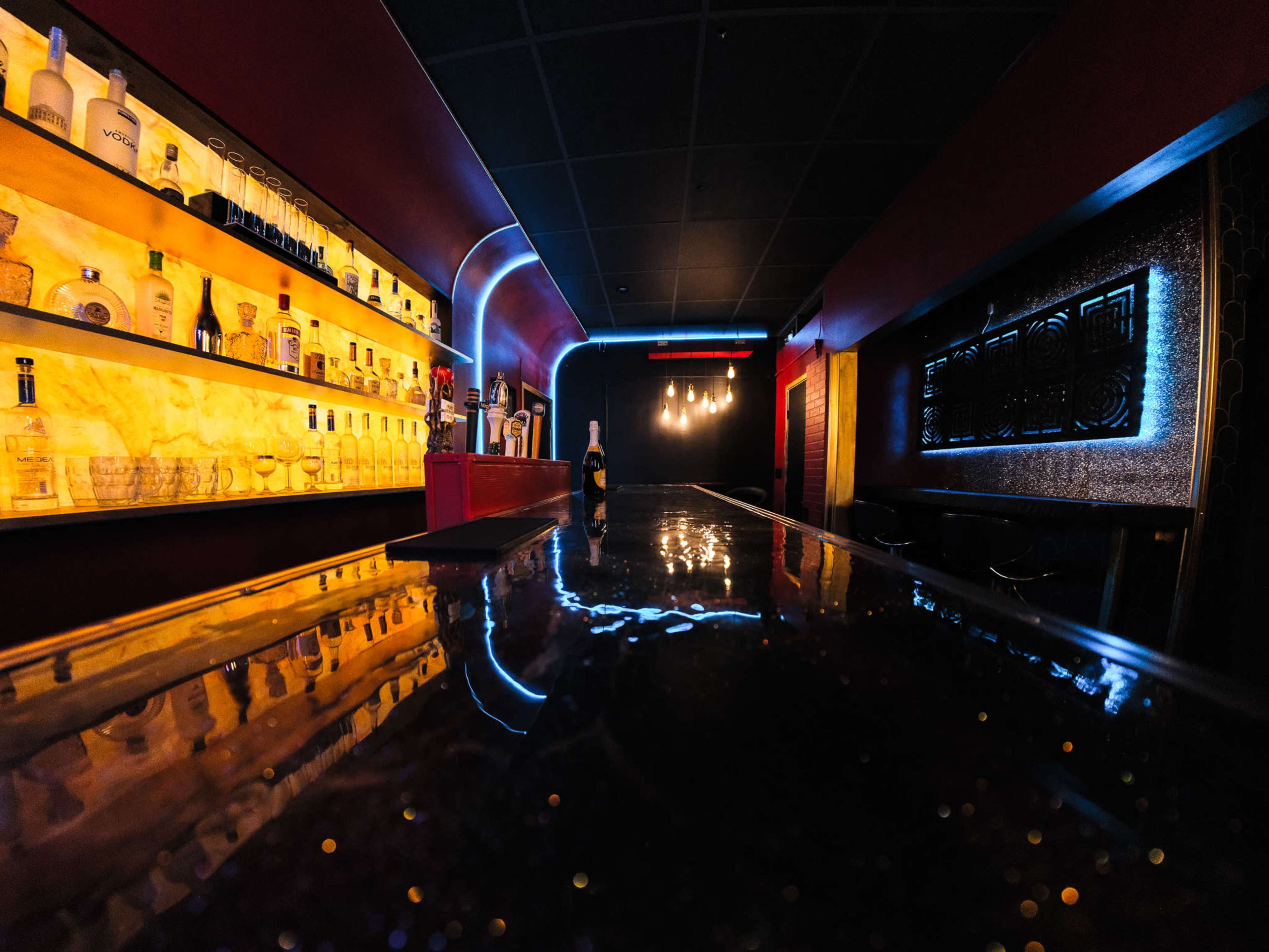 The image shows a dimly lit bar with a sleek black countertop and shelves displaying various bottles of liquor against a backdrop of illuminated, colorful walls.