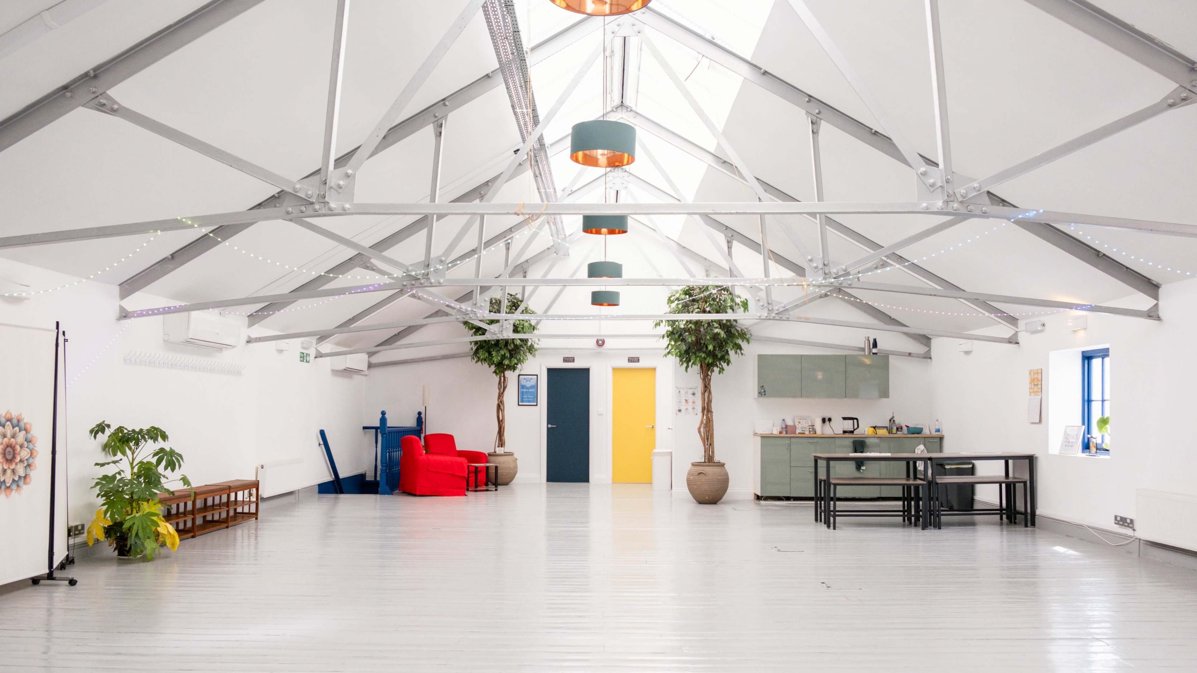 The image shows a spacious, modern interior with a high, vaulted ceiling, featuring a mix of seating areas, a kitchenette, and potted plants.
