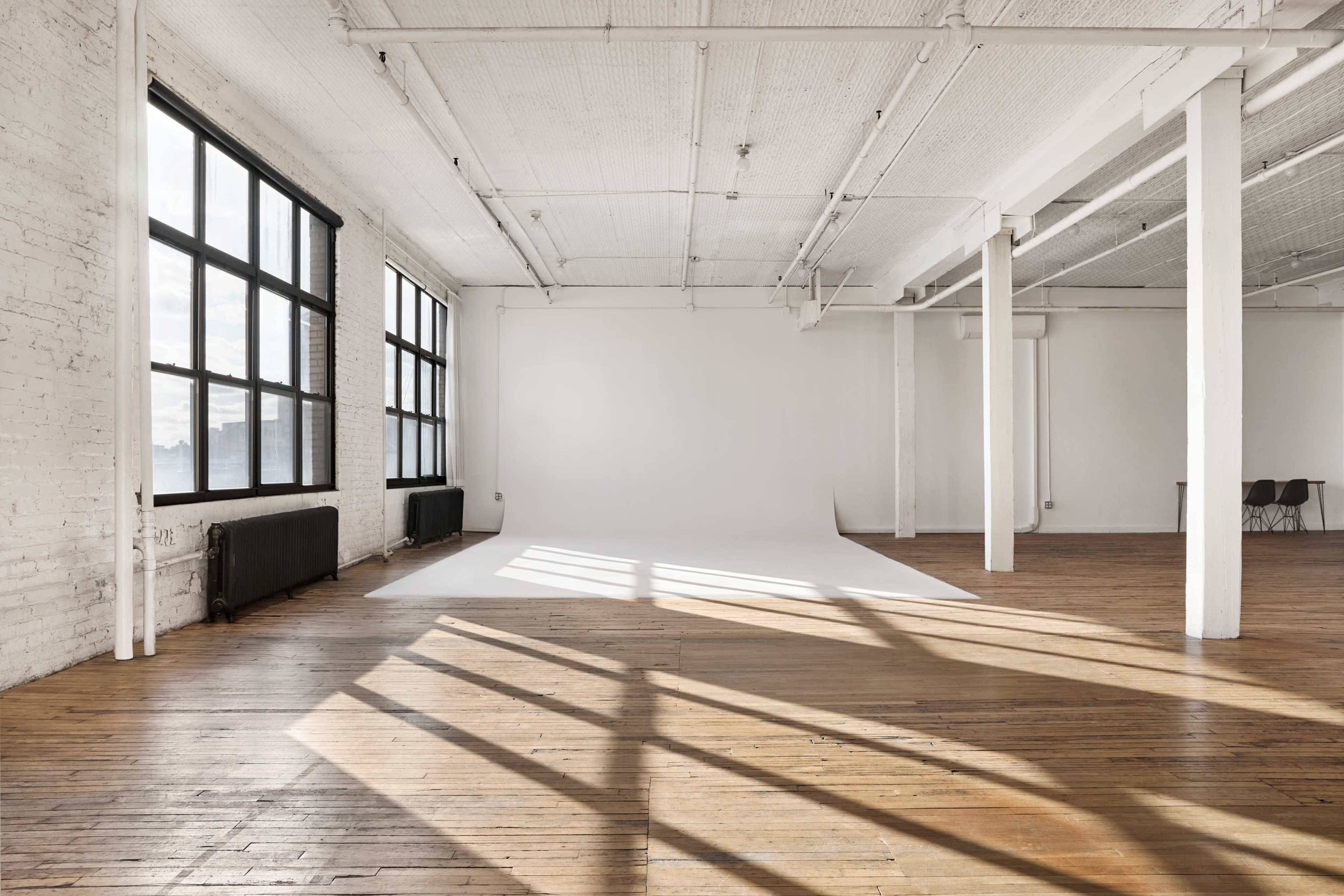 The image shows a spacious, well-lit studio with large windows, exposed brick walls, and hardwood flooring, featuring a seamless white backdrop against one wall.