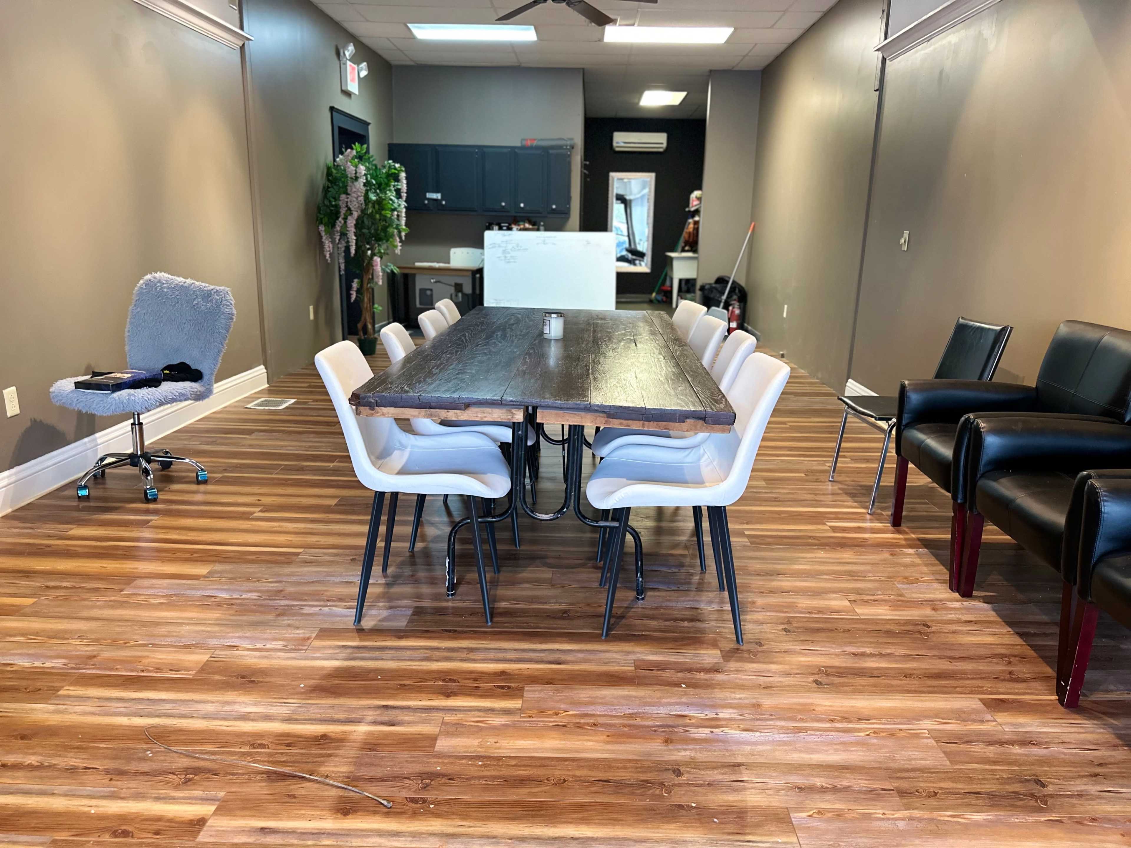 A long wooden table with white chairs is positioned in a spacious, well-lit room featuring wooden flooring and gray walls.