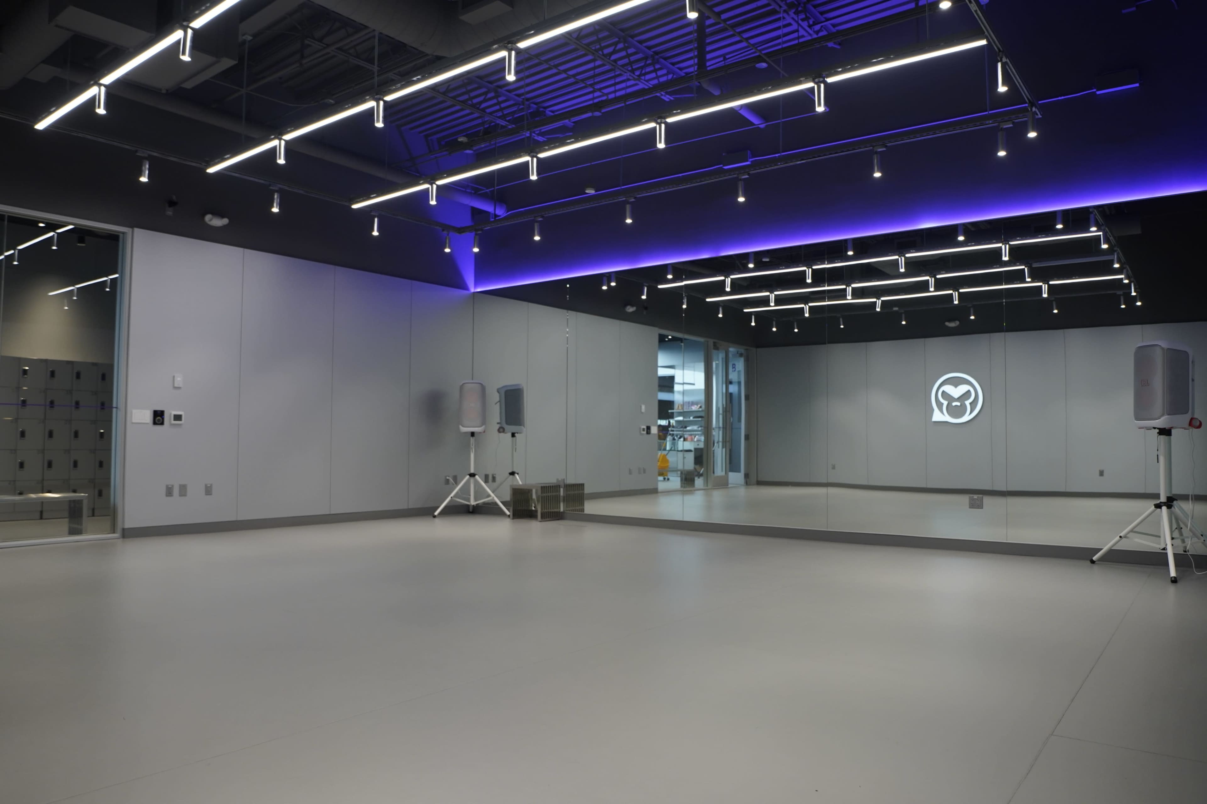 The image shows a modern studio space featuring a large mirrored wall, sound equipment on stands, and bright overhead lighting in a minimalist design.