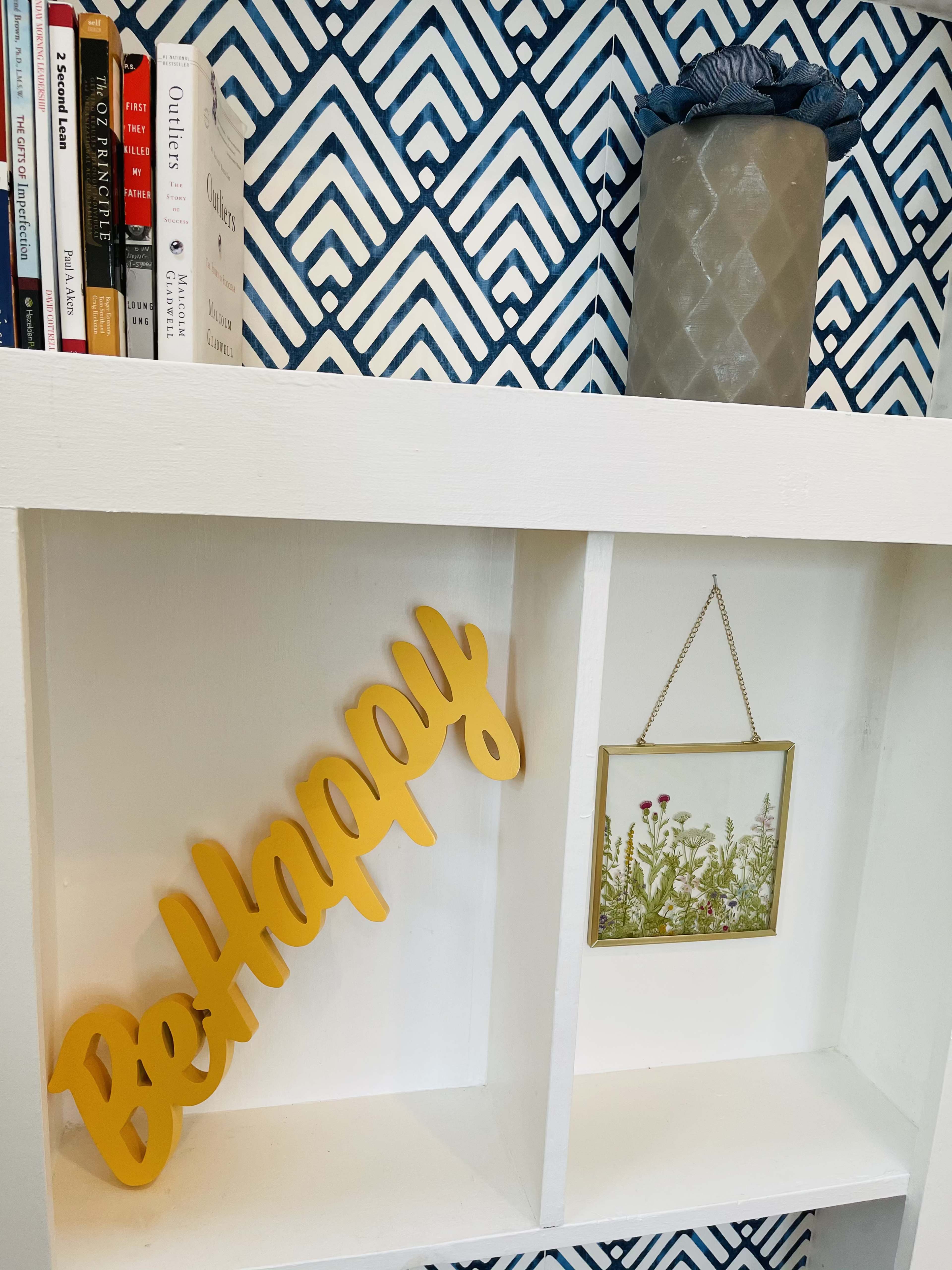 A shelf with a yellow "Be Happy" sign, a framed picture of flowers, and books lined on a blue patterned wall.