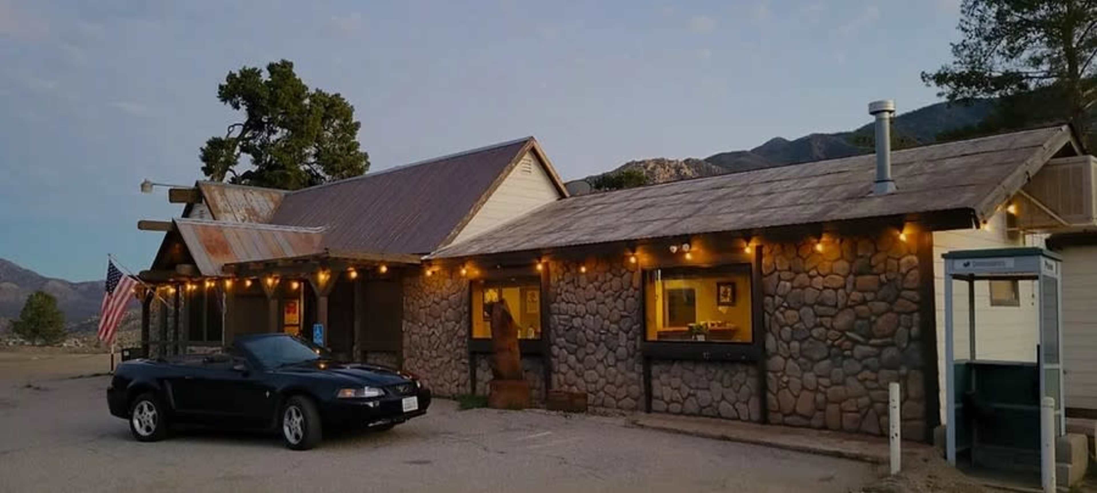 A stone building with a metal roof and string lights is shown alongside a parked black convertible car and an American flag.