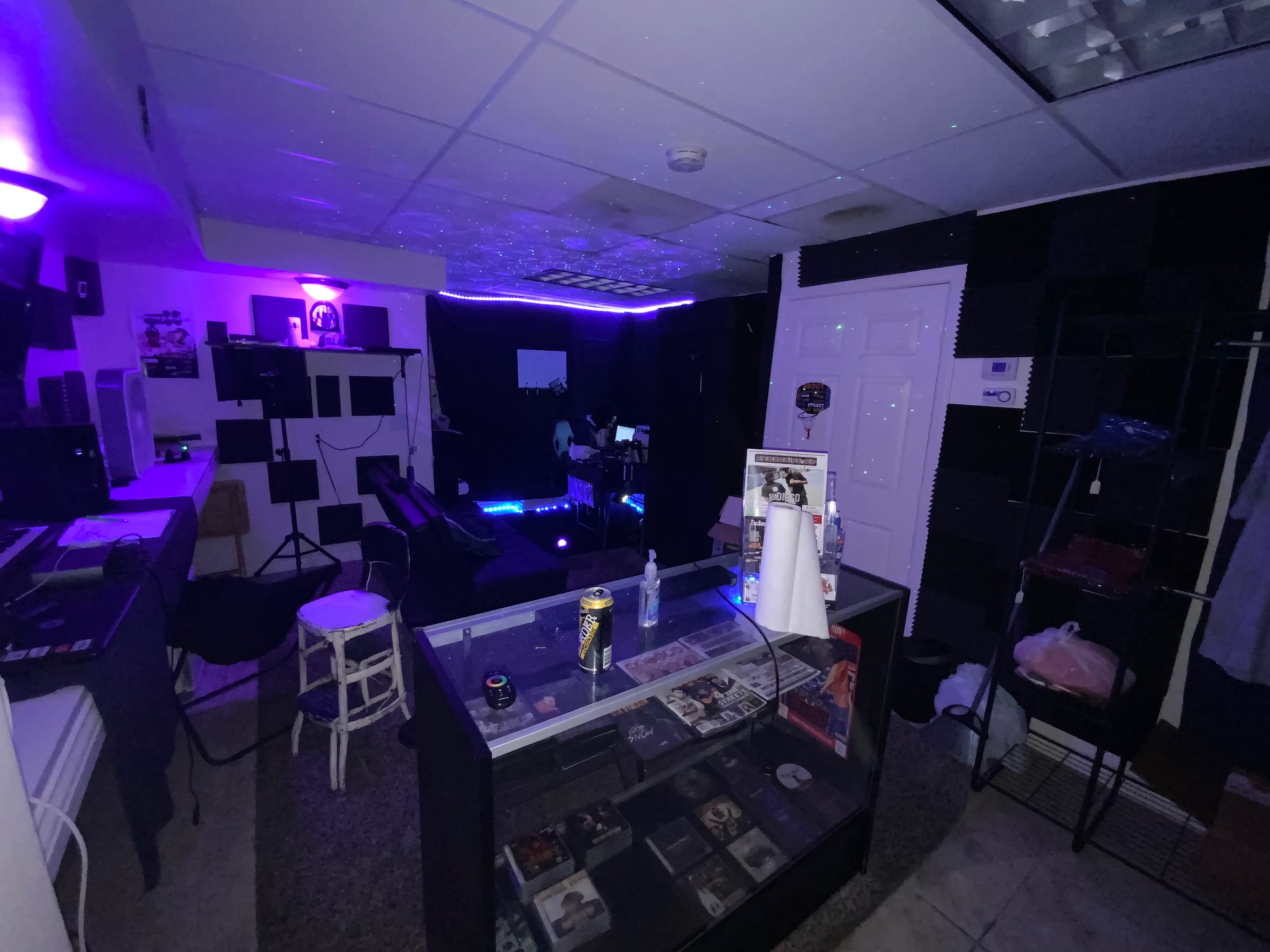 A dimly lit recording studio with purple lighting, featuring a mixing console, shelves of CDs, and soundproofing panels on the walls.