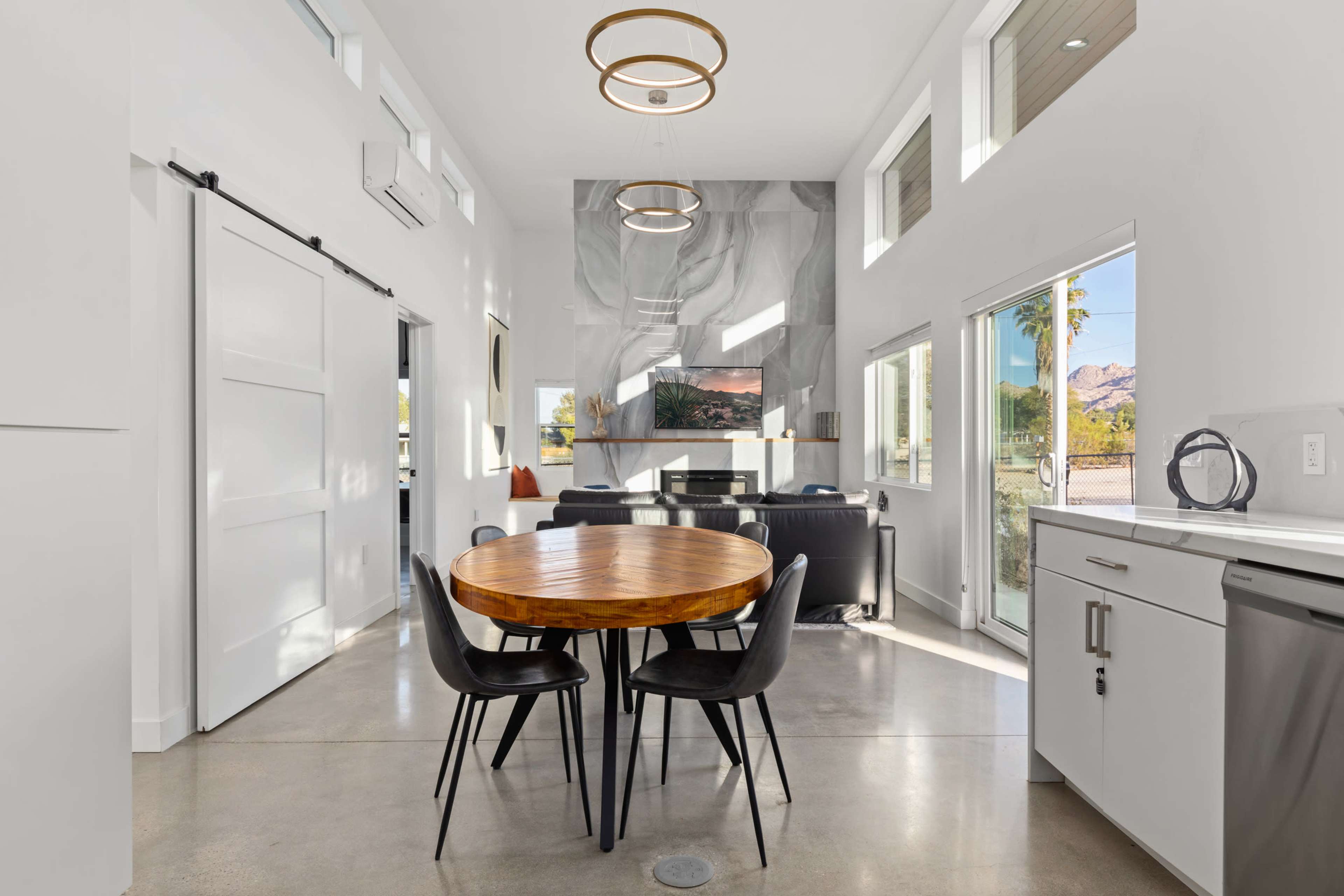 A modern dining area features a round wooden table surrounded by black chairs, with large windows and a marble accent wall in the background.