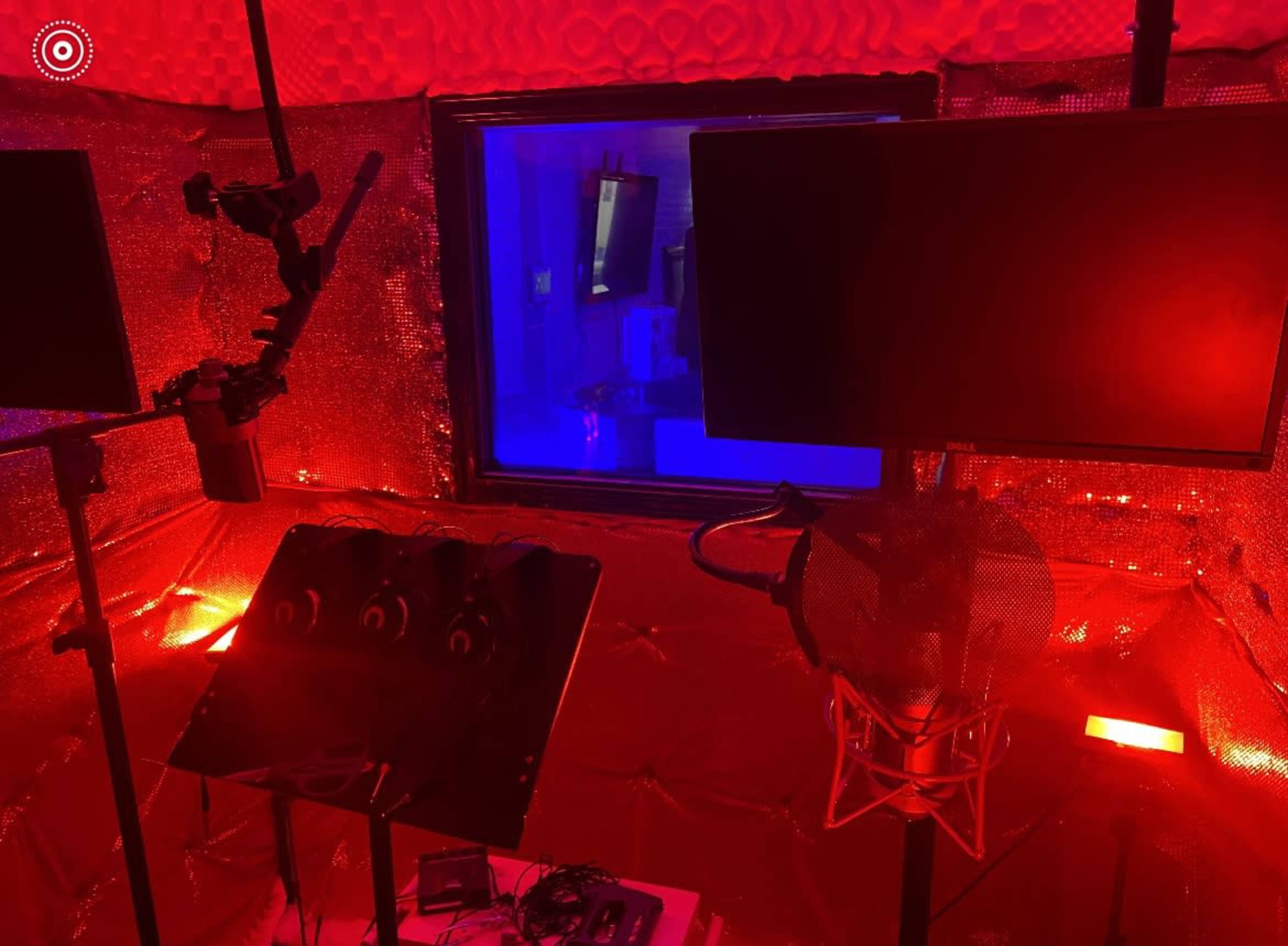 A recording studio with a soundproof booth, microphones, and monitors illuminated in red and blue light.
