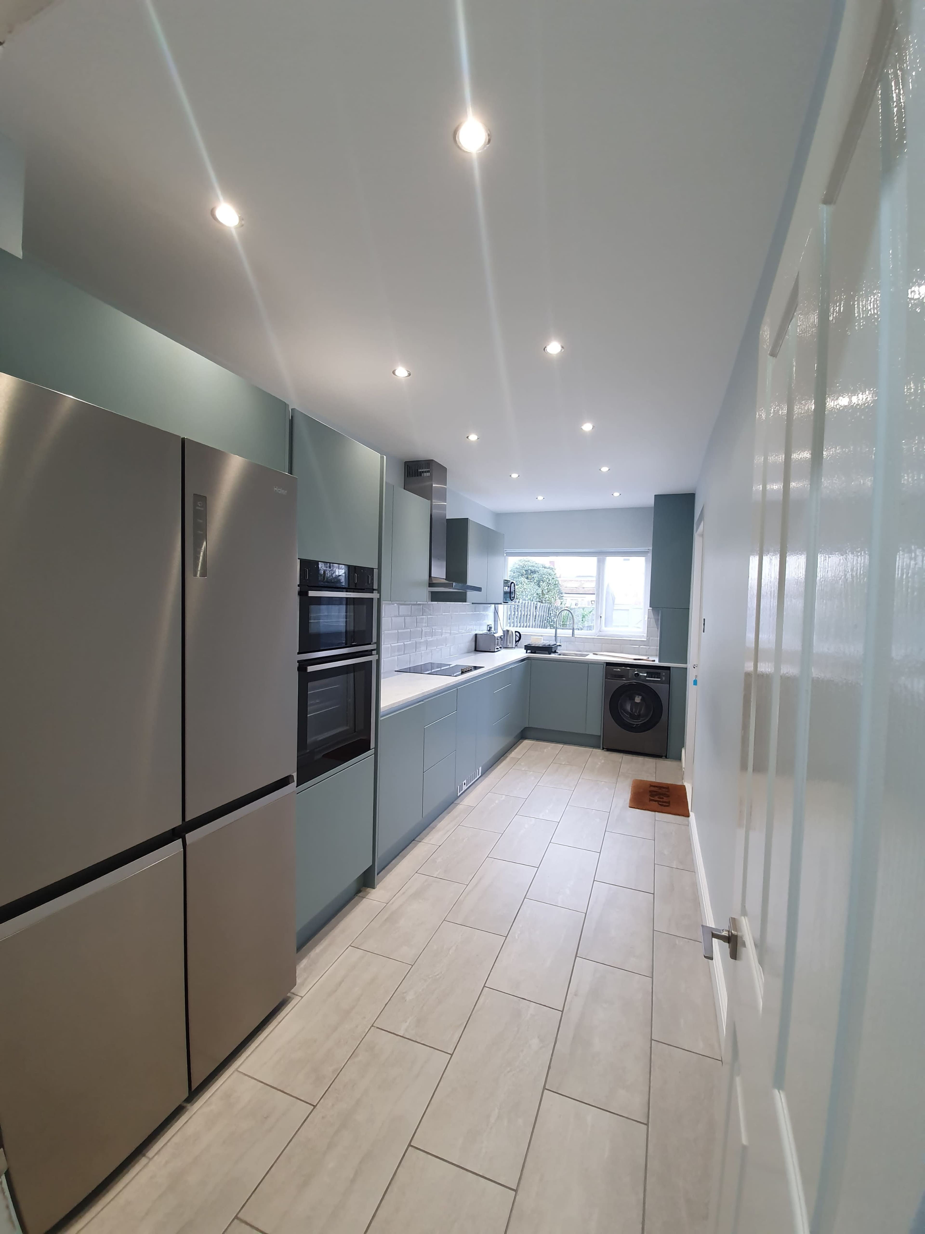 A modern kitchen with light blue cabinetry, stainless steel appliances, and a tiled floor, leading to a window view.