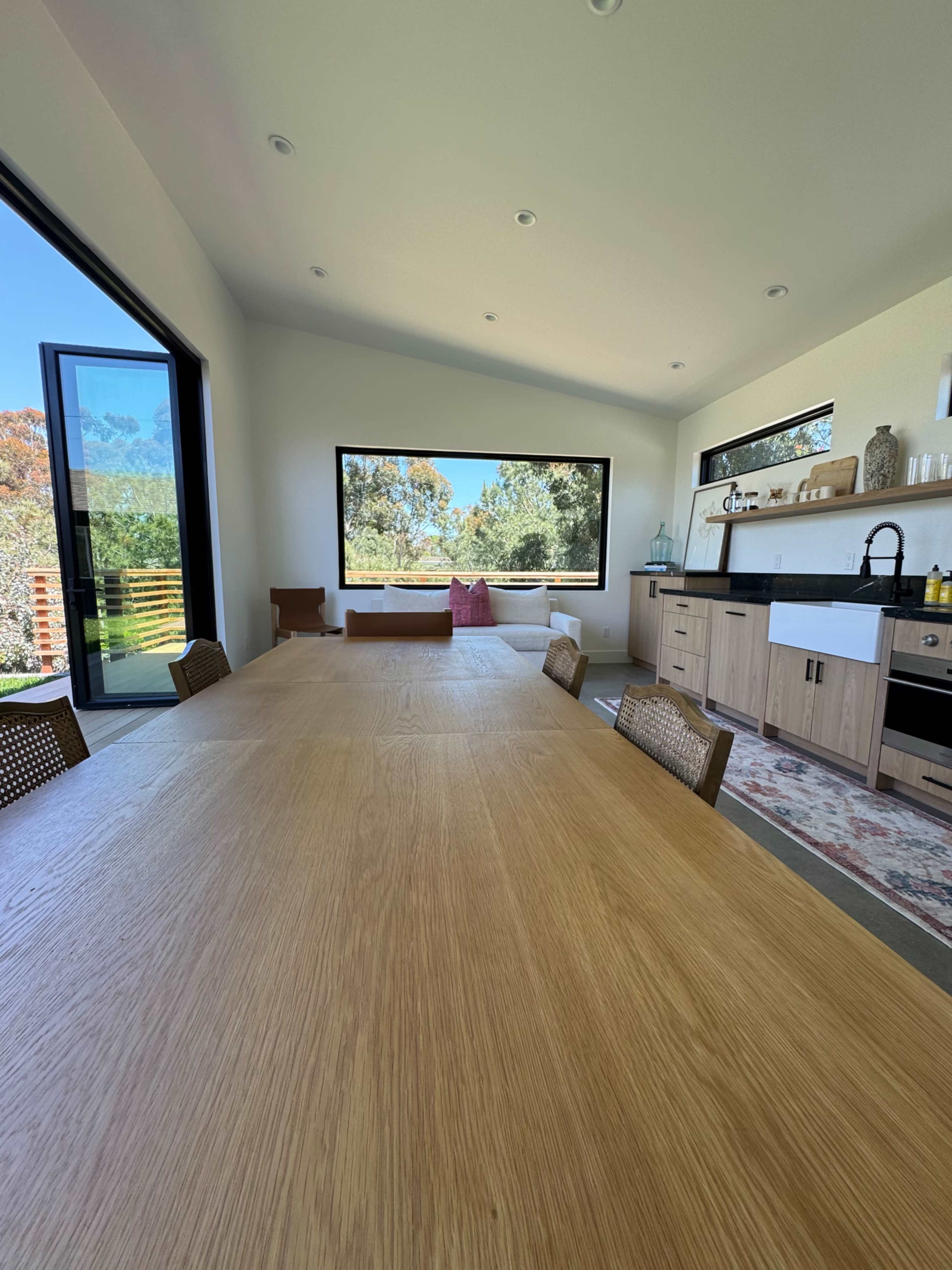 A spacious dining area features a long wooden table and a kitchen counter with large windows overlooking a green landscape.