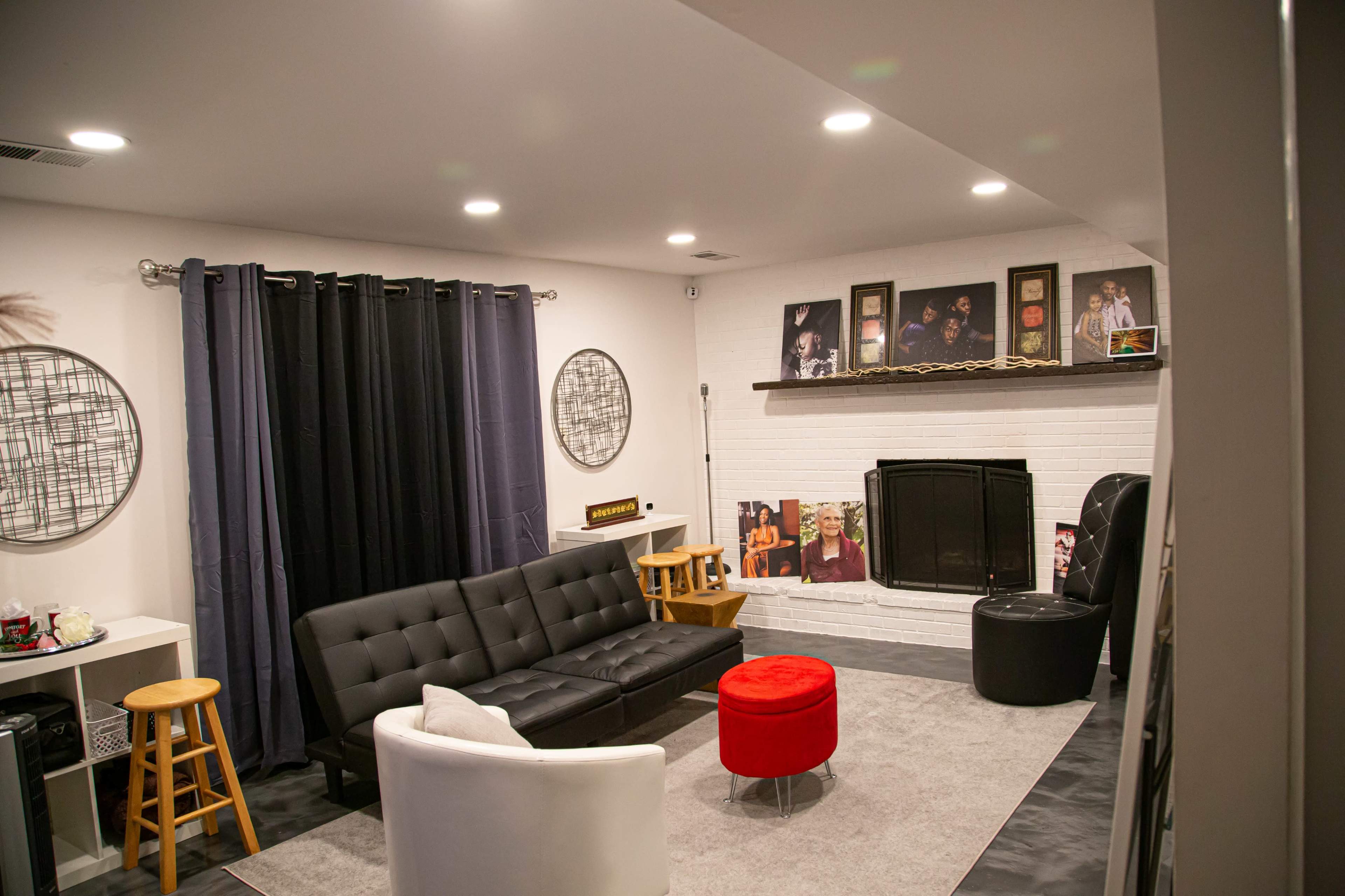 A modern living room features a black sectional sofa, a white armchair, a red accent table, and framed photographs on the walls, with a fireplace and gray area rug.
