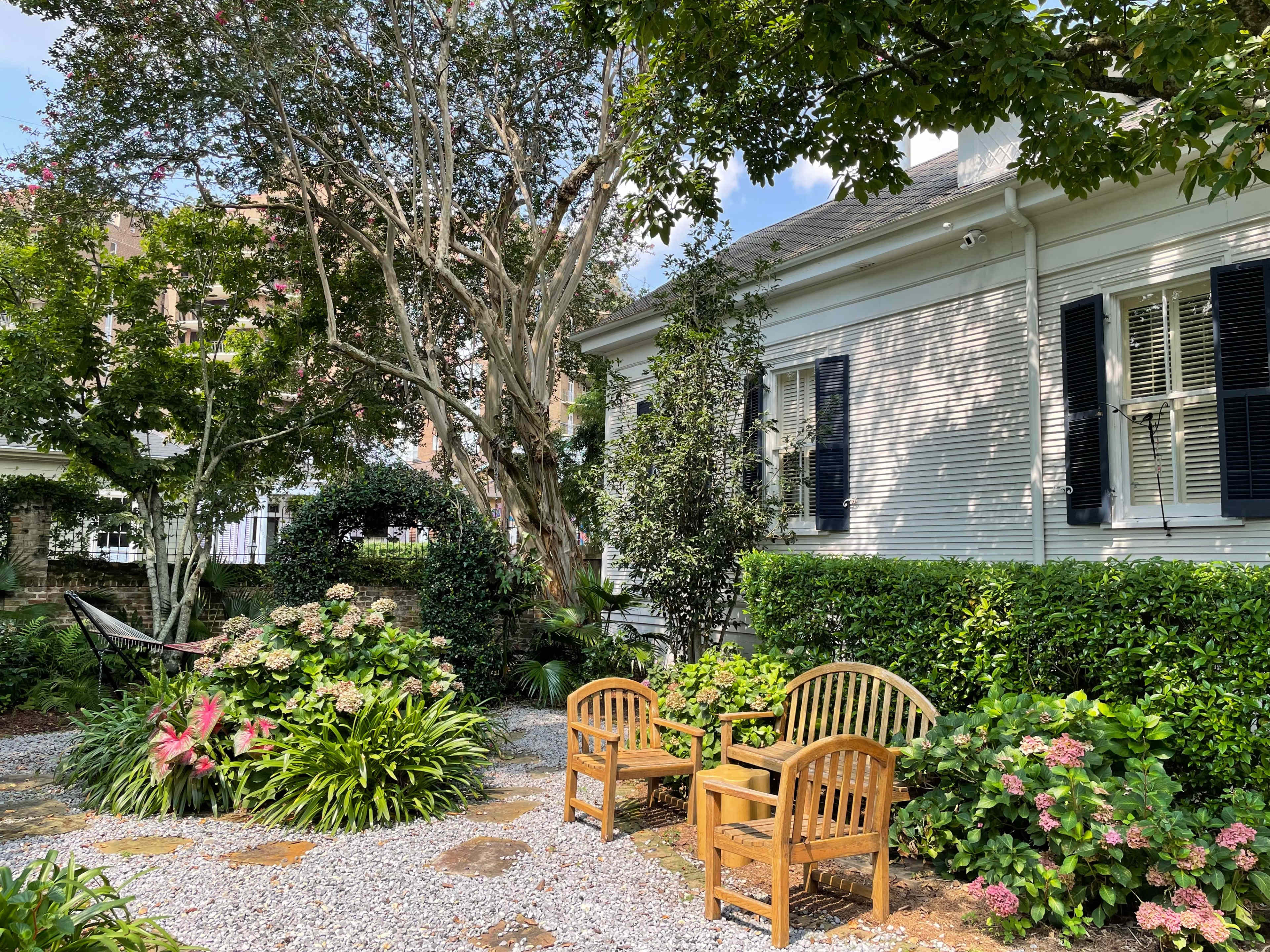 A garden features two wooden chairs surrounded by lush greenery and flowering plants beside a white house.