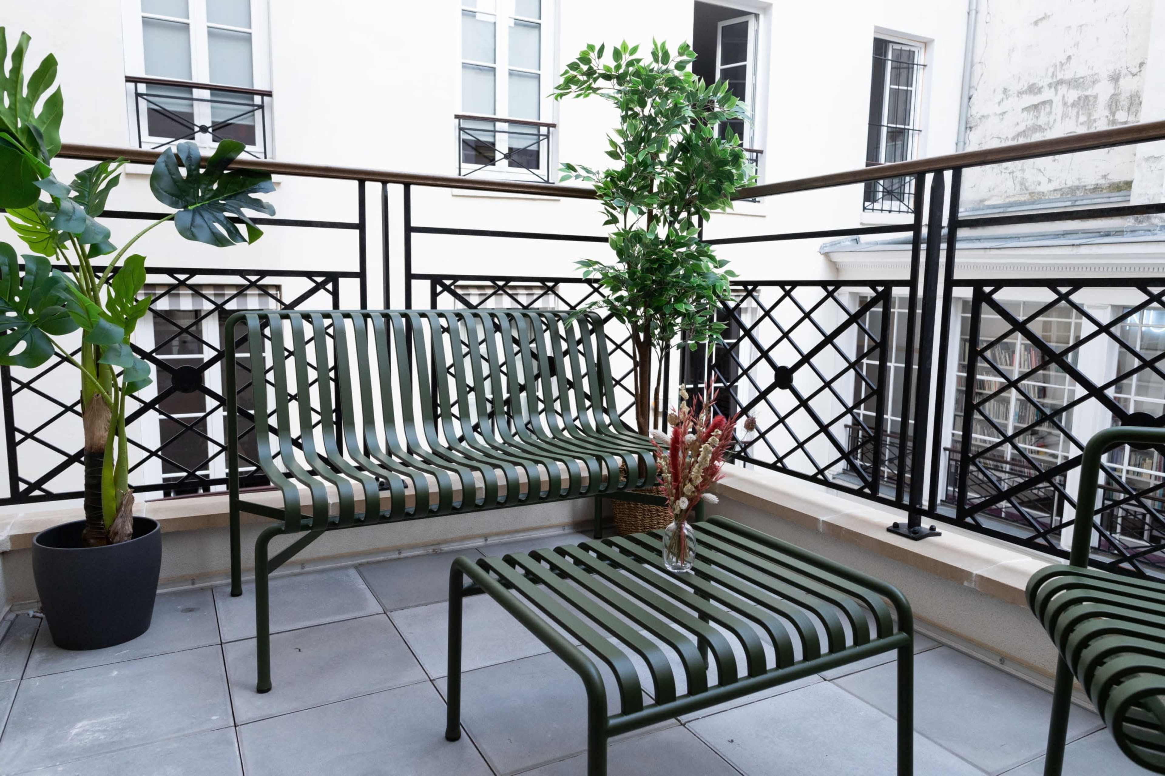 A small balcony features green striped metal furniture, including a sofa and a coffee table, alongside potted plants and a decorative arrangement.