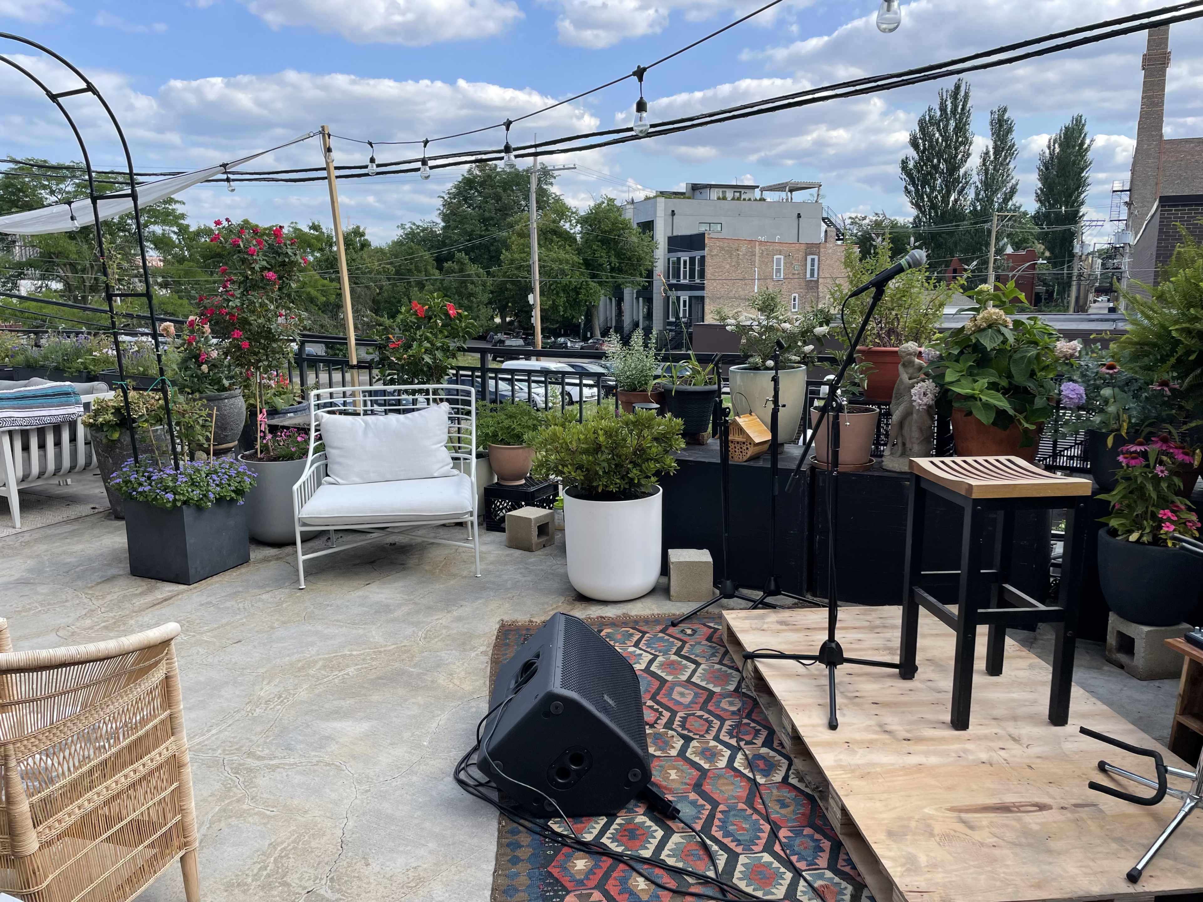 A rooftop space features potted plants, a microphone setup on a wooden platform, and seating arrangements with a scenic backdrop.