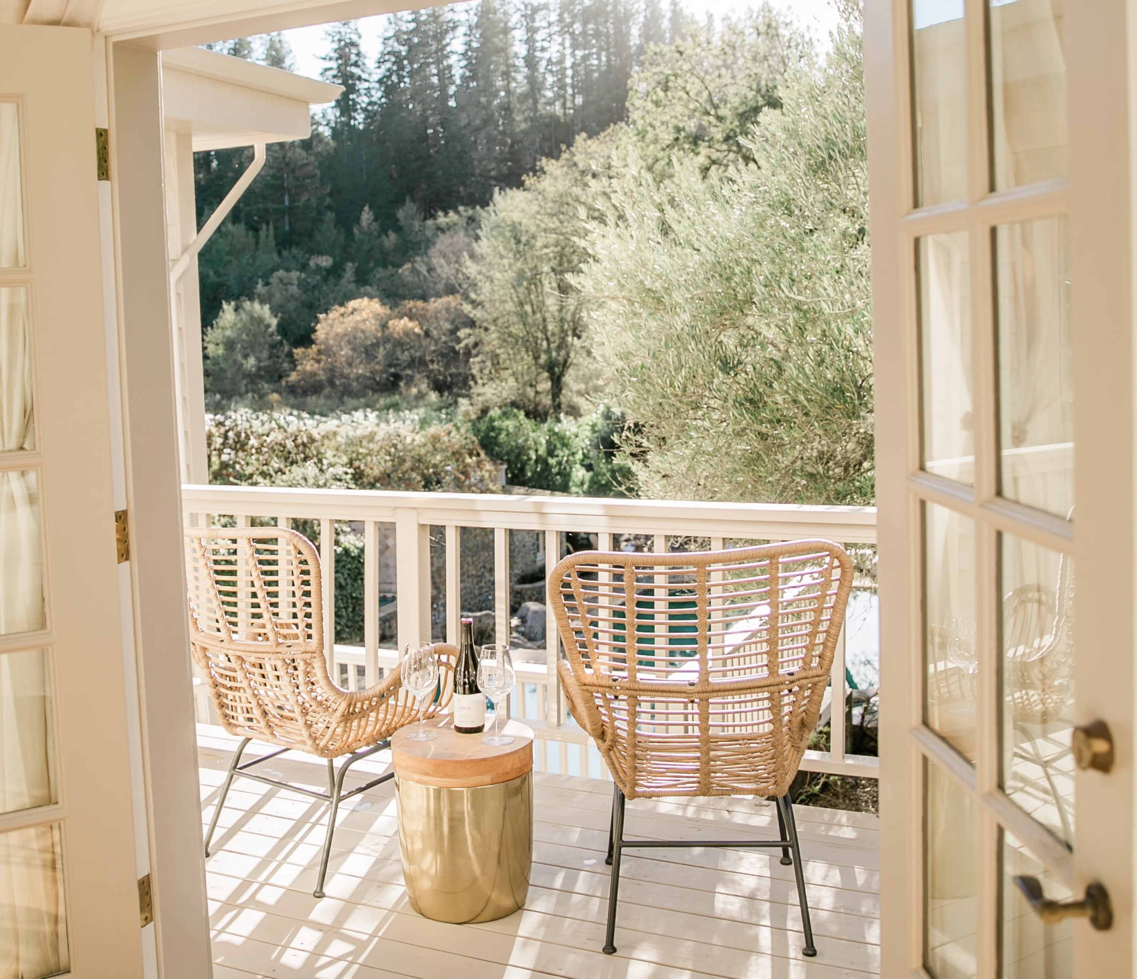 A balcony features two rattan chairs beside a small metallic table, overlooking a lush green landscape.