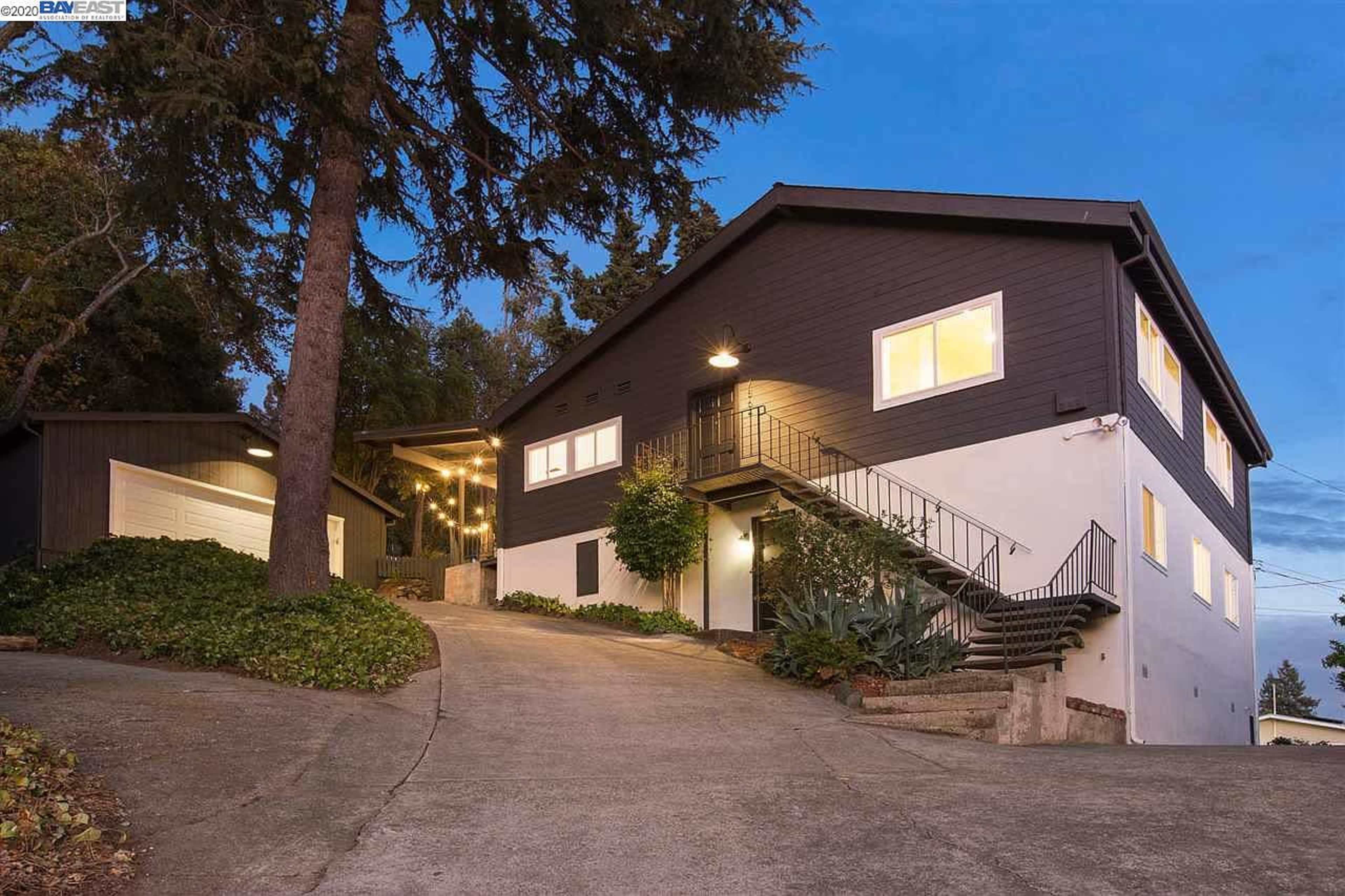 A two-story house situated on a sloped driveway, with a garage on the left and multiple windows illuminated in the evening light.