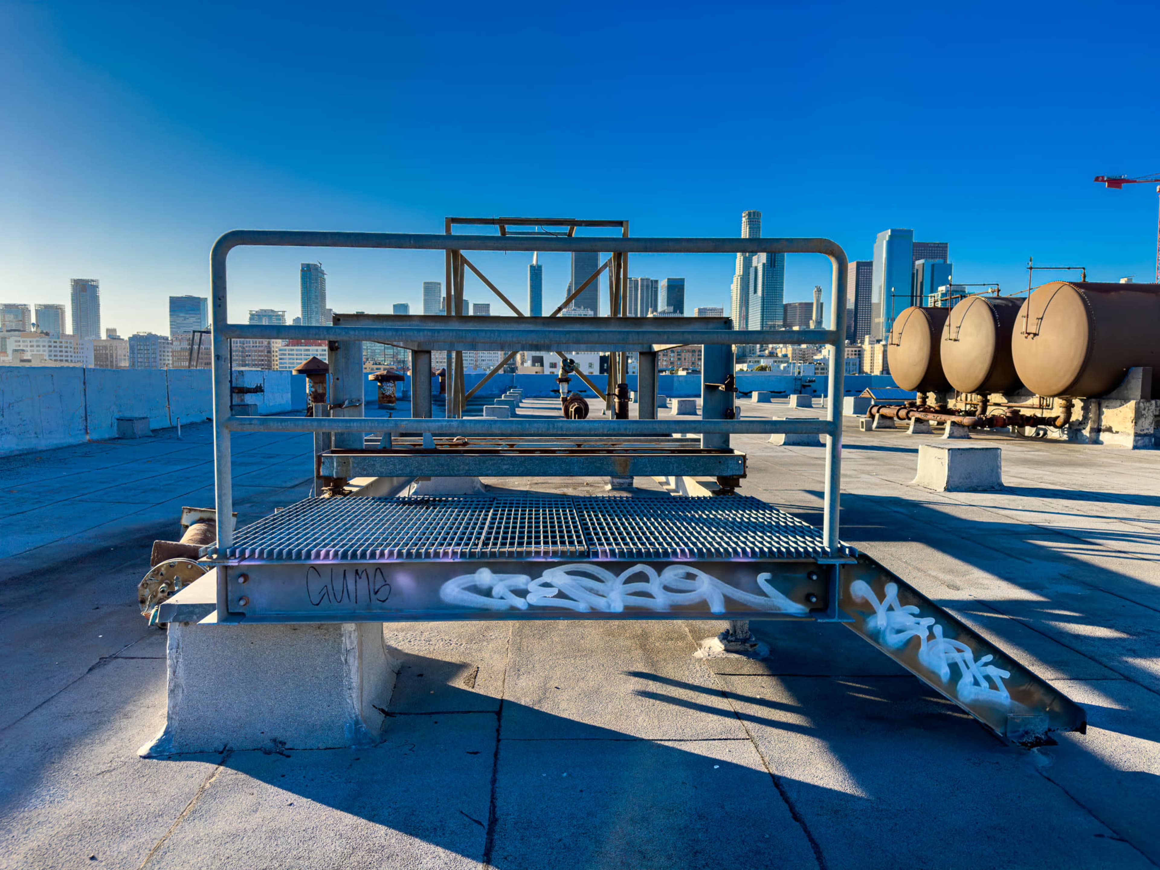 A metal platform with graffiti is positioned on a rooftop, overlooking a city skyline with tall buildings and storage tanks in the background.