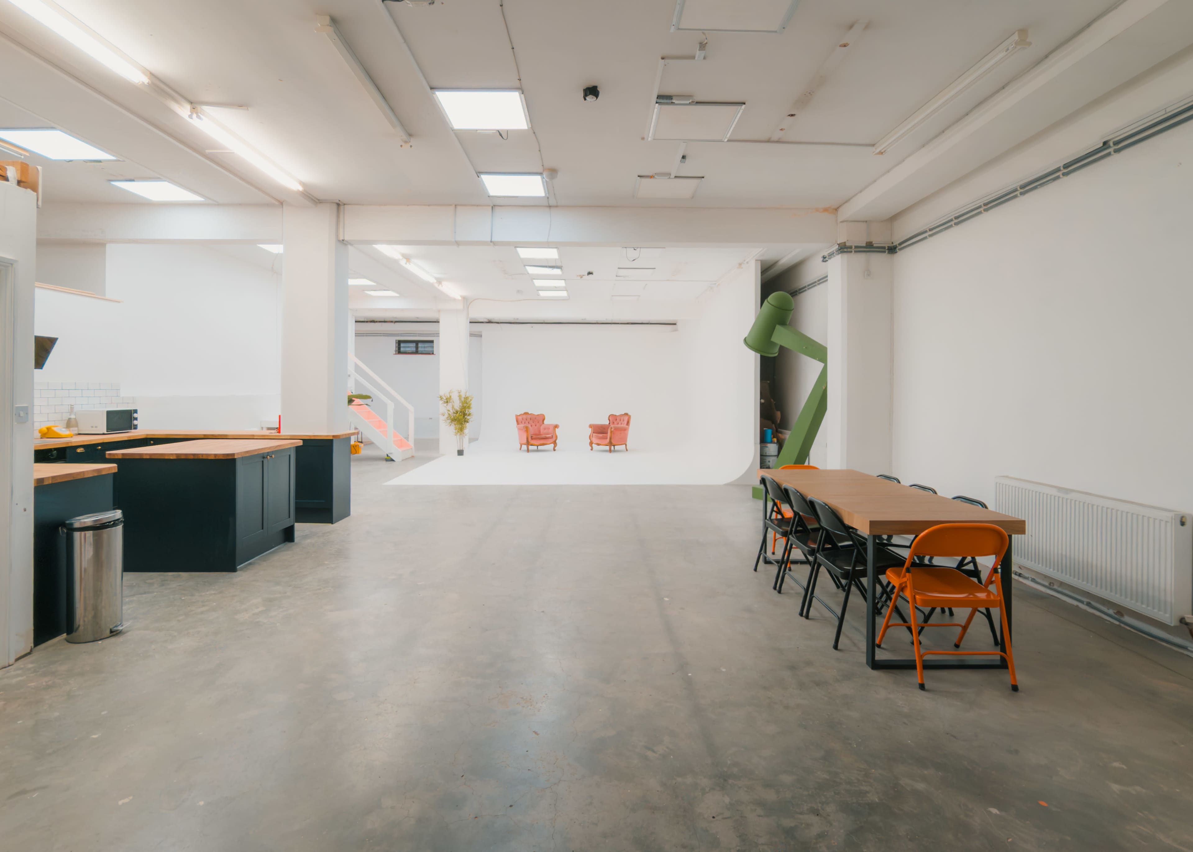 A spacious, well-lit studio featuring a kitchen area on the left, a large open space in the center, and two red chairs positioned against a blank backdrop at the far end.