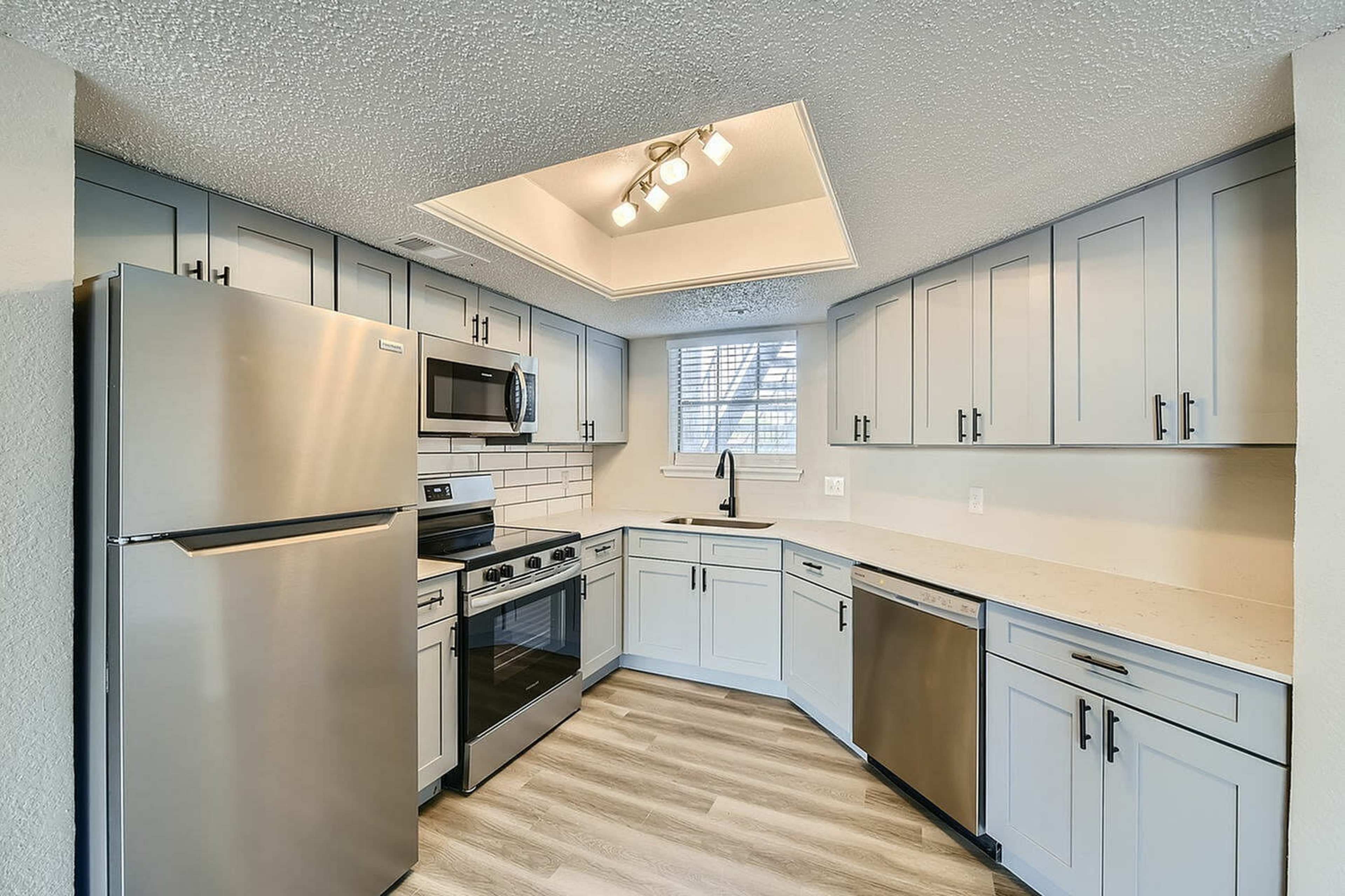 The image shows a modern kitchen with light gray cabinets, stainless steel appliances, and a large window providing natural light.