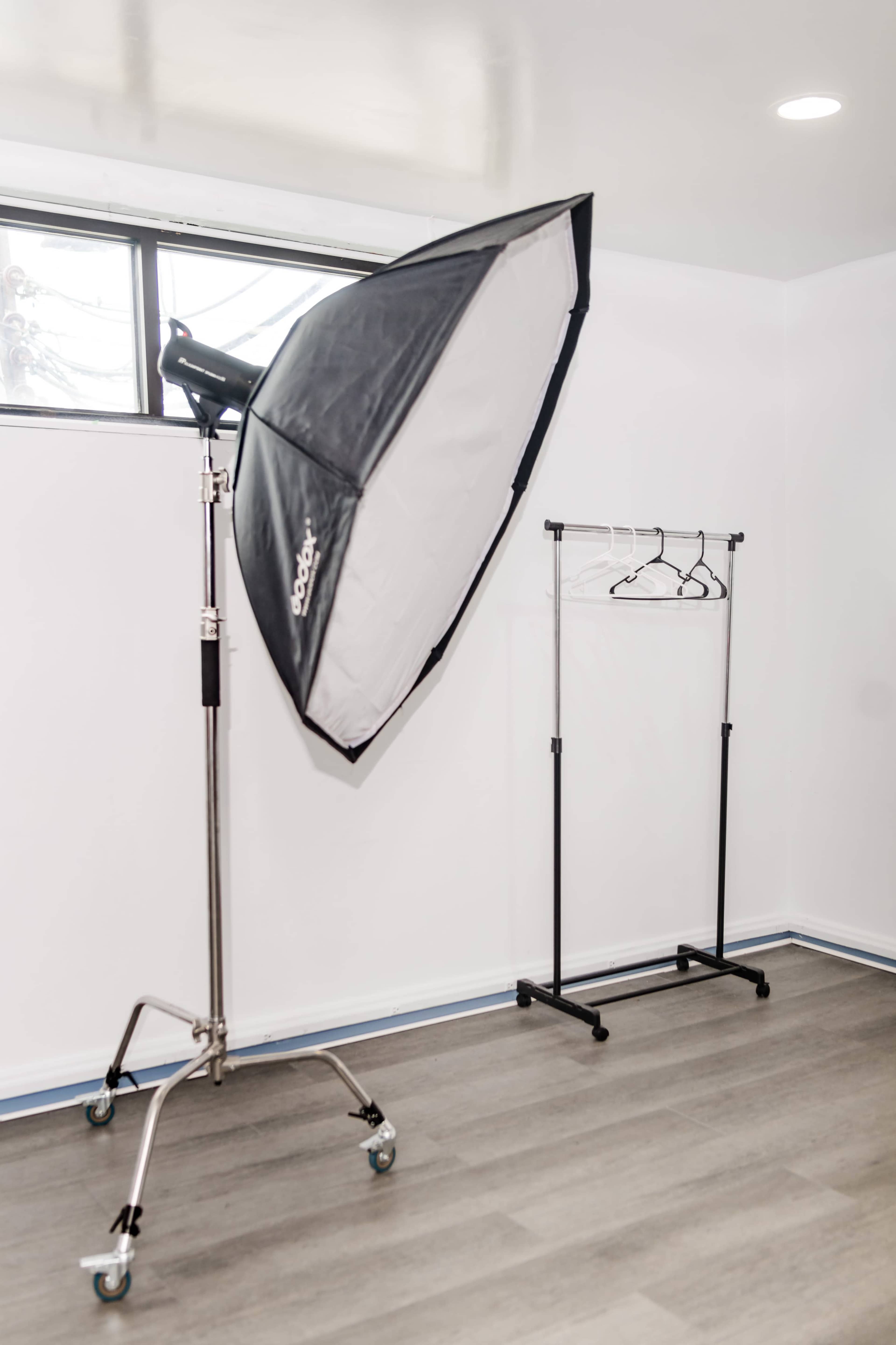 A photography studio with a large softbox light on a stand and a clothing rack against a white wall.