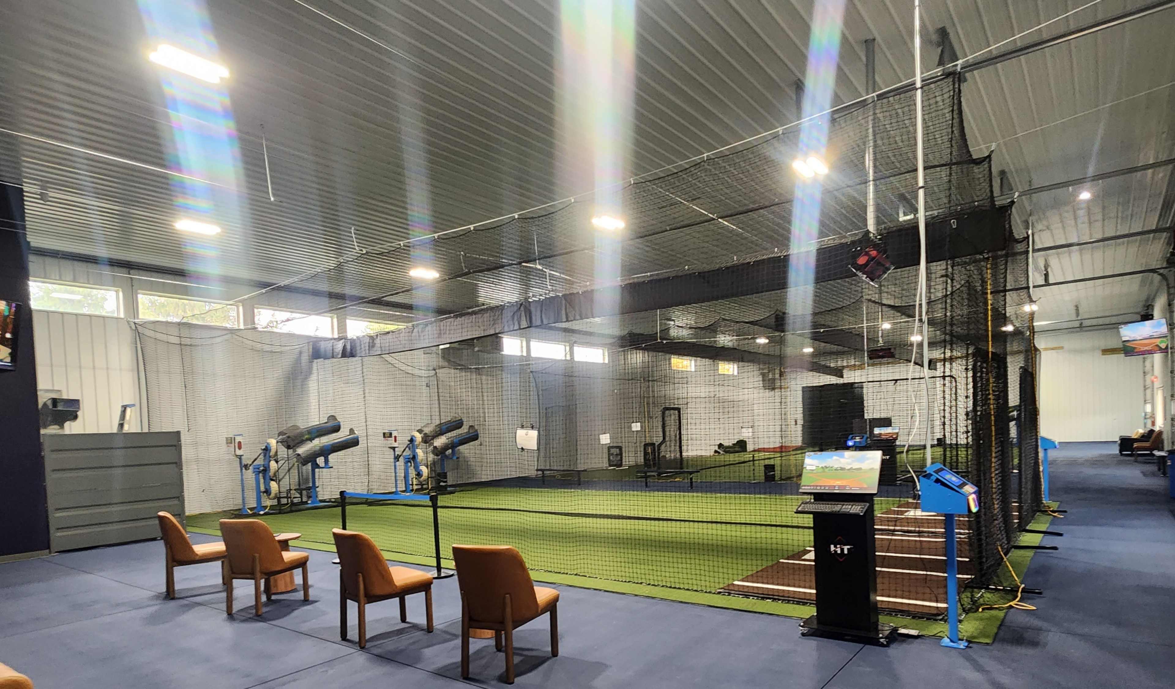 The image shows an indoor batting facility featuring multiple batting cages, artificial turf, and a seating area with chairs.