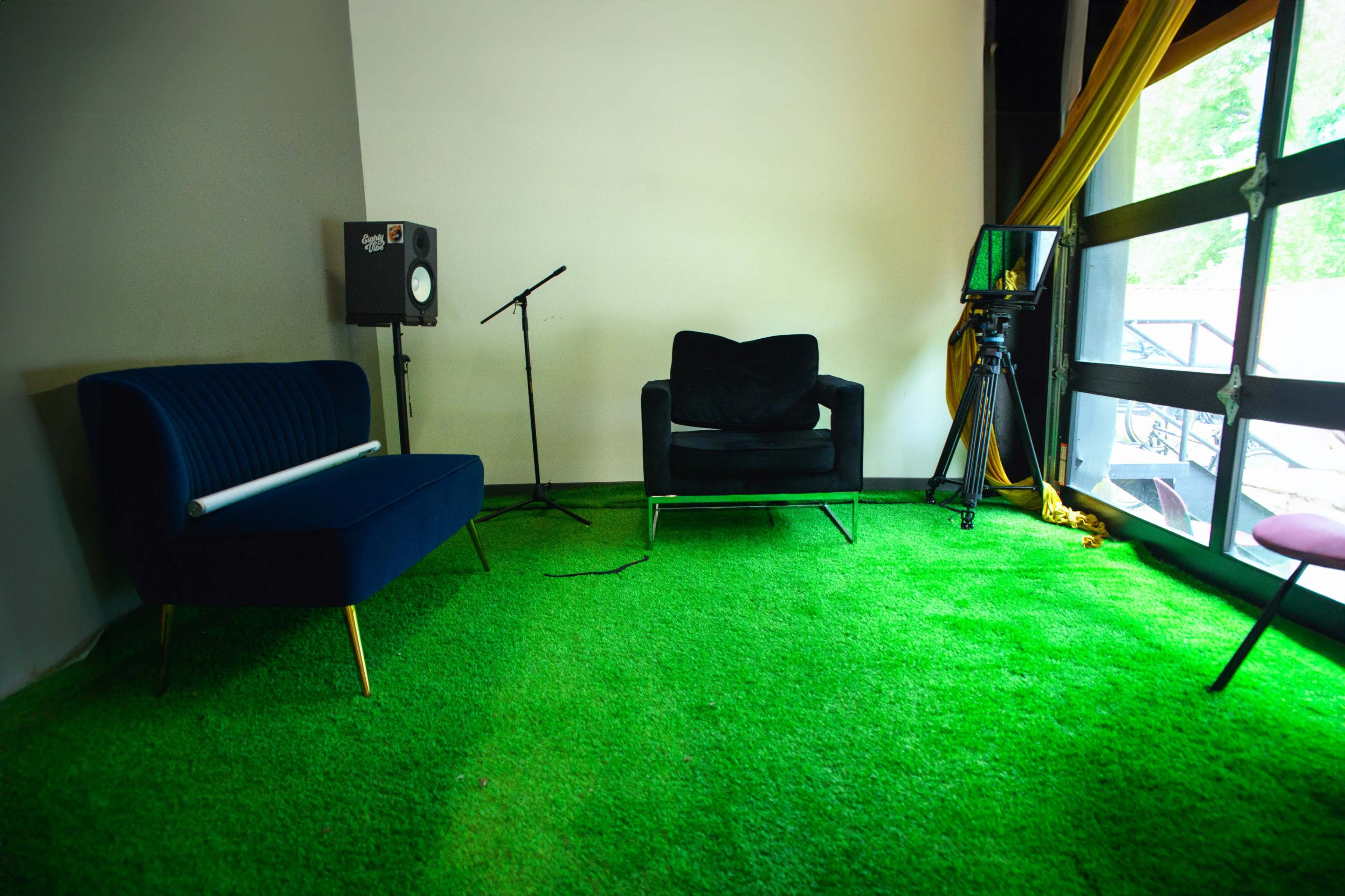 The image shows a brightly lit room featuring two chairs on green carpet, a microphone on a stand, and a camera set up near a window.