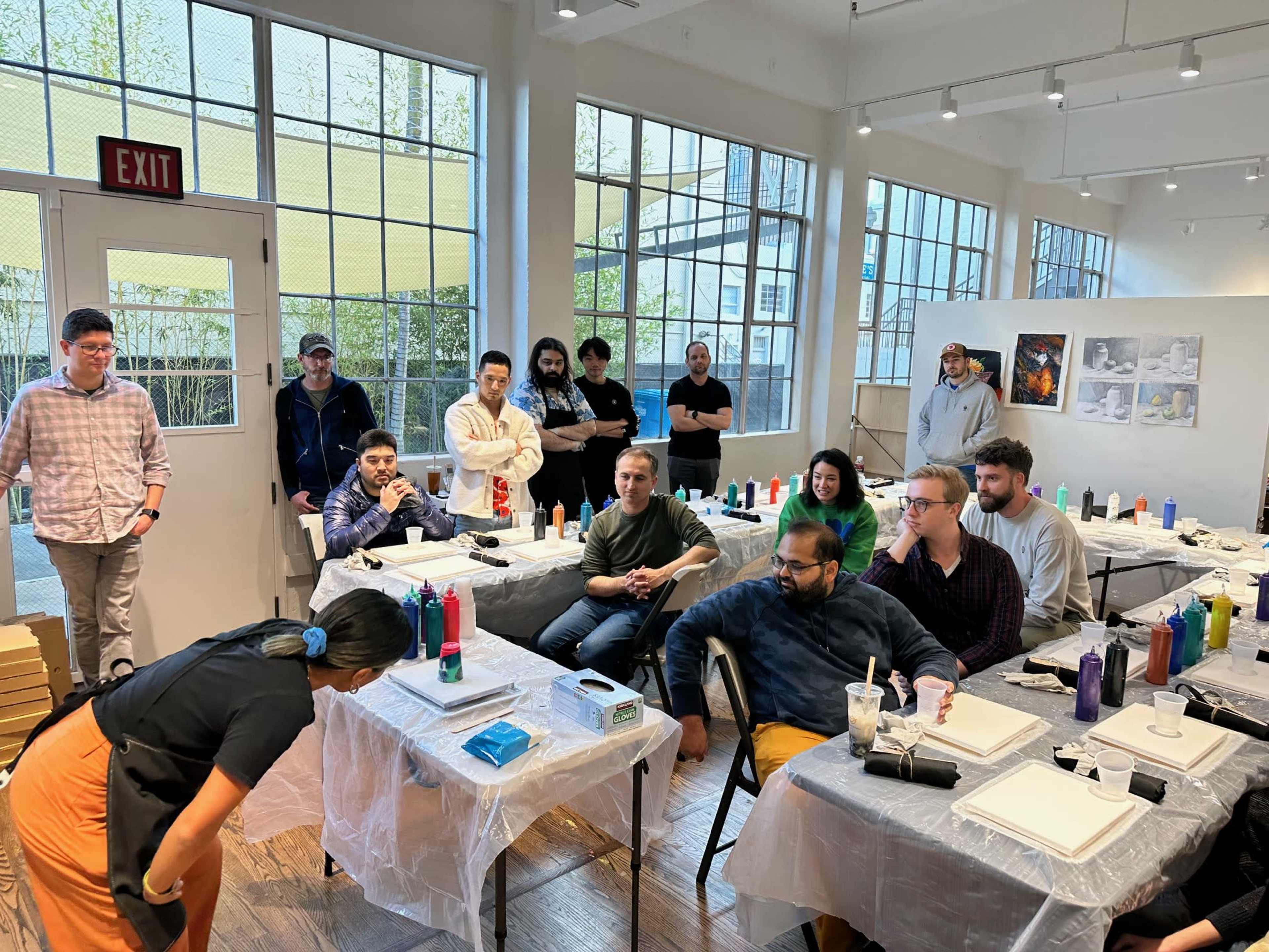 A group of individuals is gathered in a well-lit workshop space, observing a demonstration at tables covered with plastic sheets.