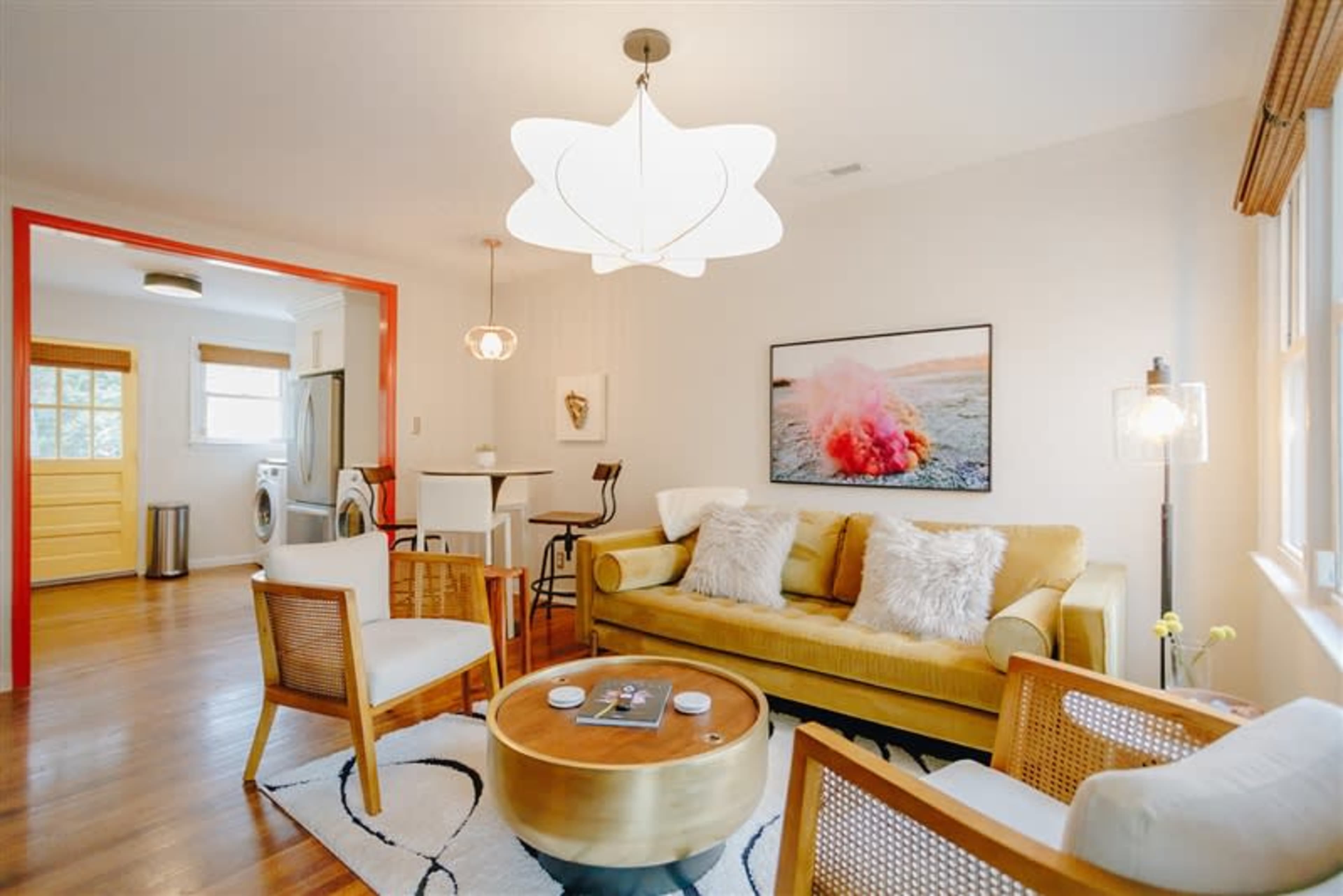 A modern living room features a yellow sofa, a round coffee table, and a large artwork on the wall, with an adjoining kitchen visible in the background.