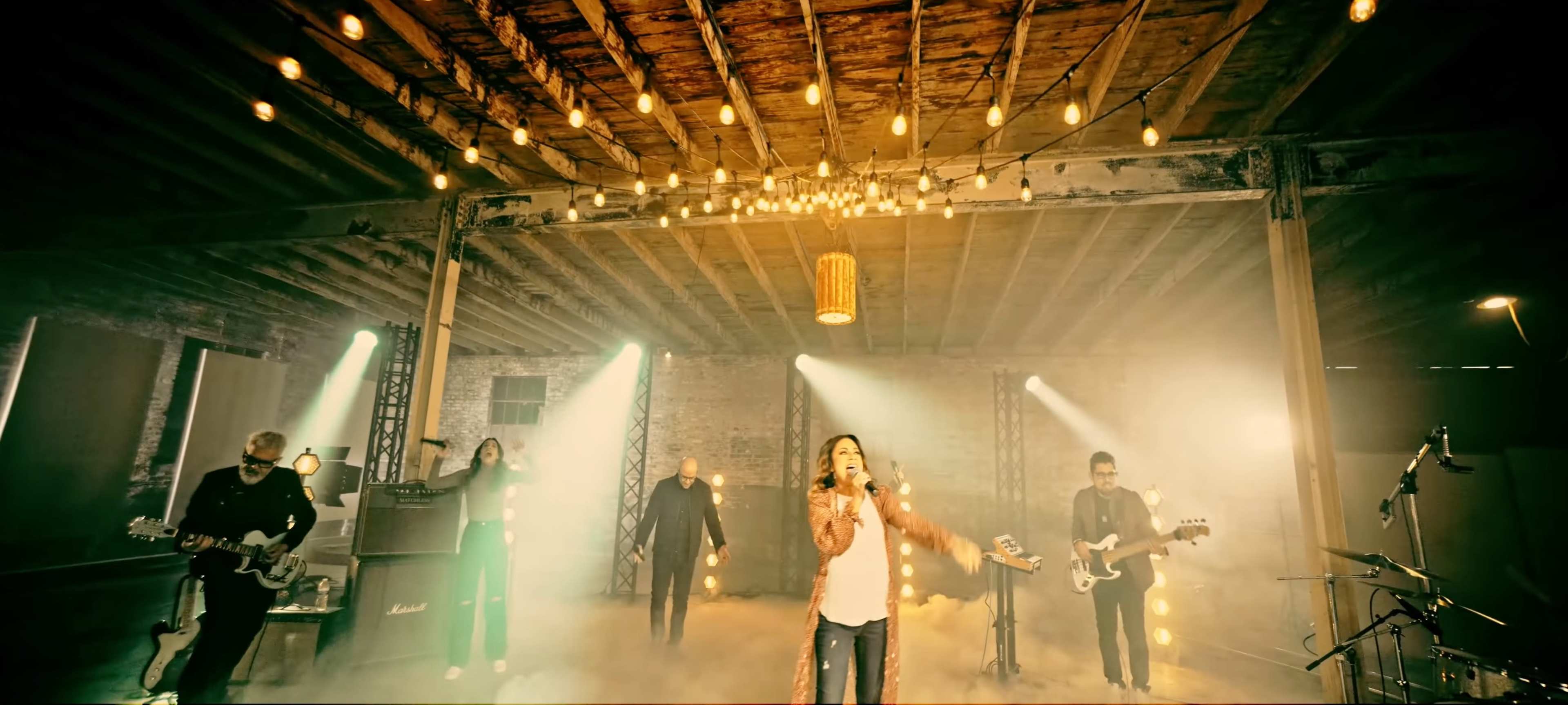 A band performs on a stage with string lights and a smoky atmosphere in a rustic setting.