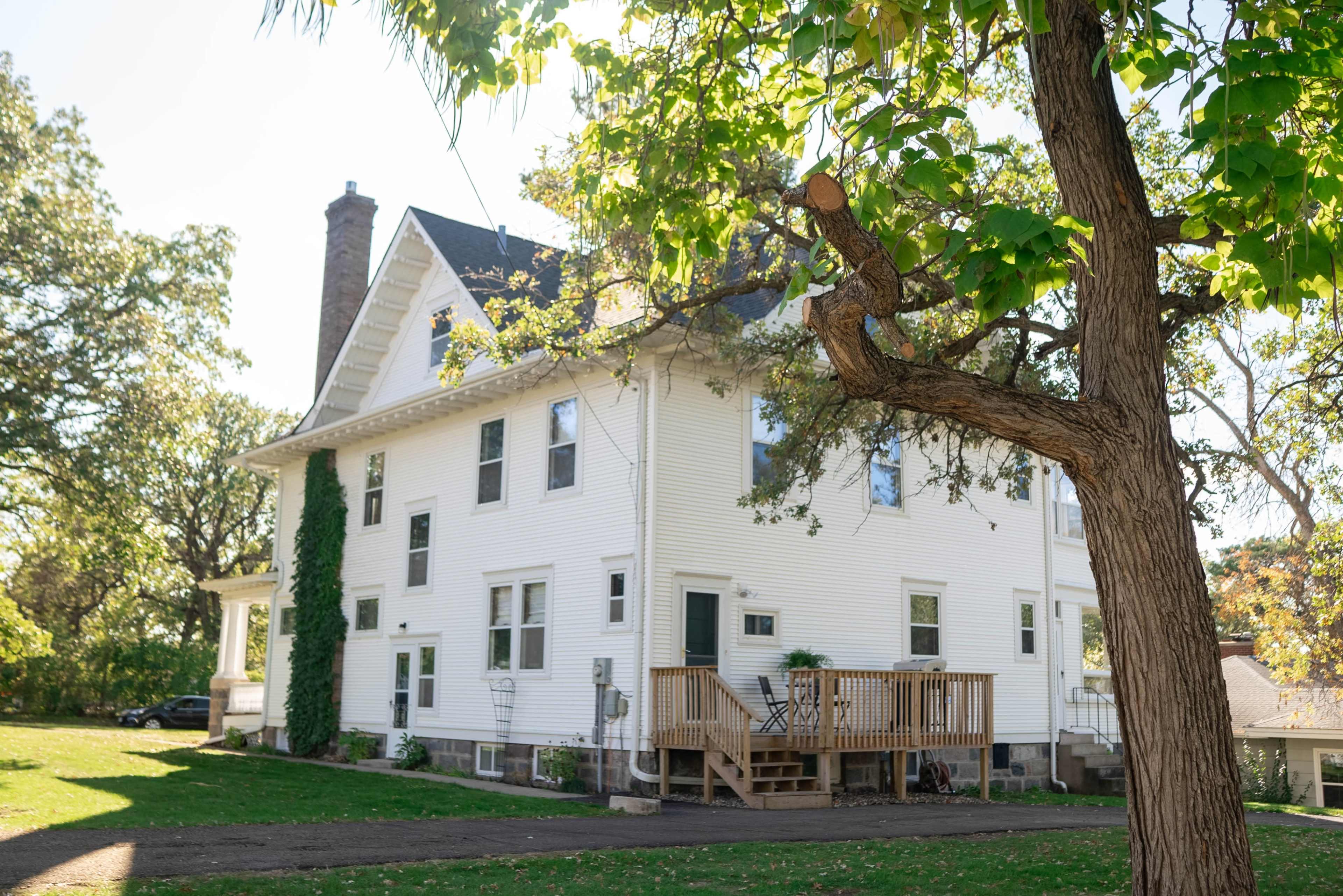 A large white house with a wooden deck is surrounded by trees in a grassy area.
