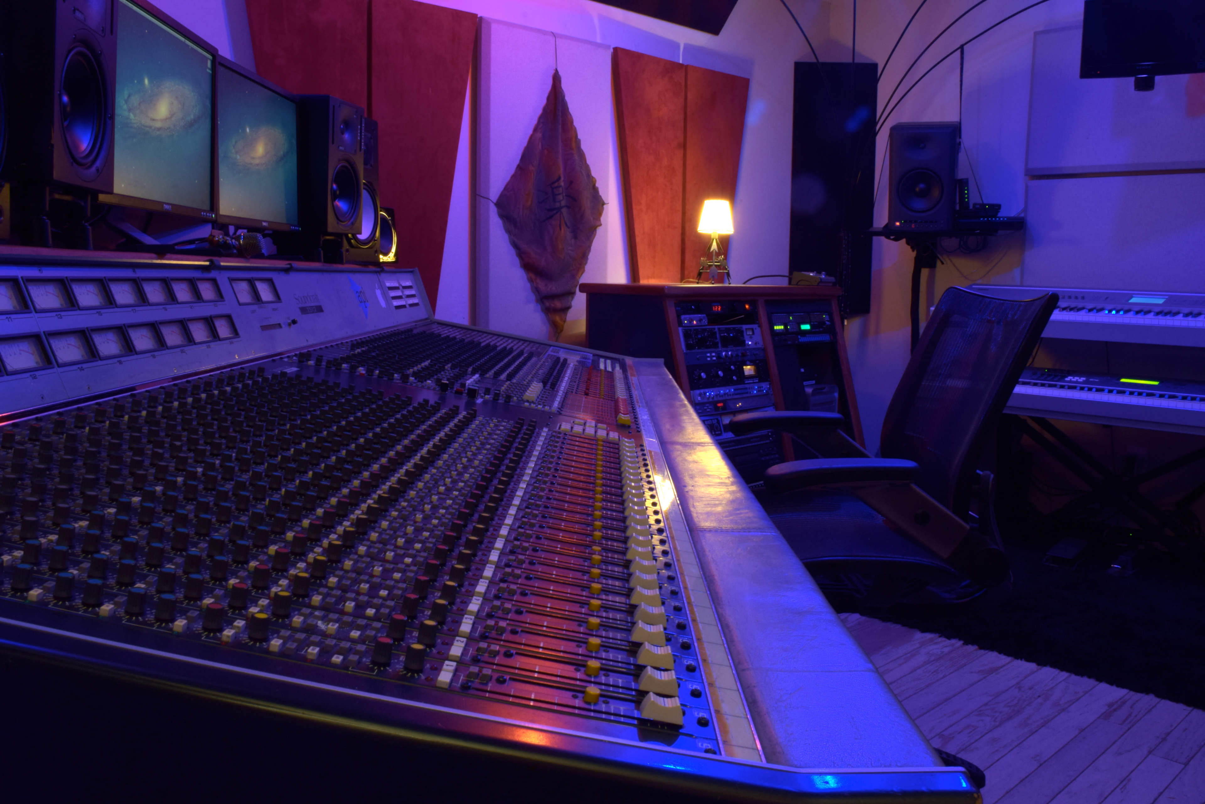 The image shows a recording studio with a large mixing console in the foreground, flanked by computer monitors and various audio equipment.