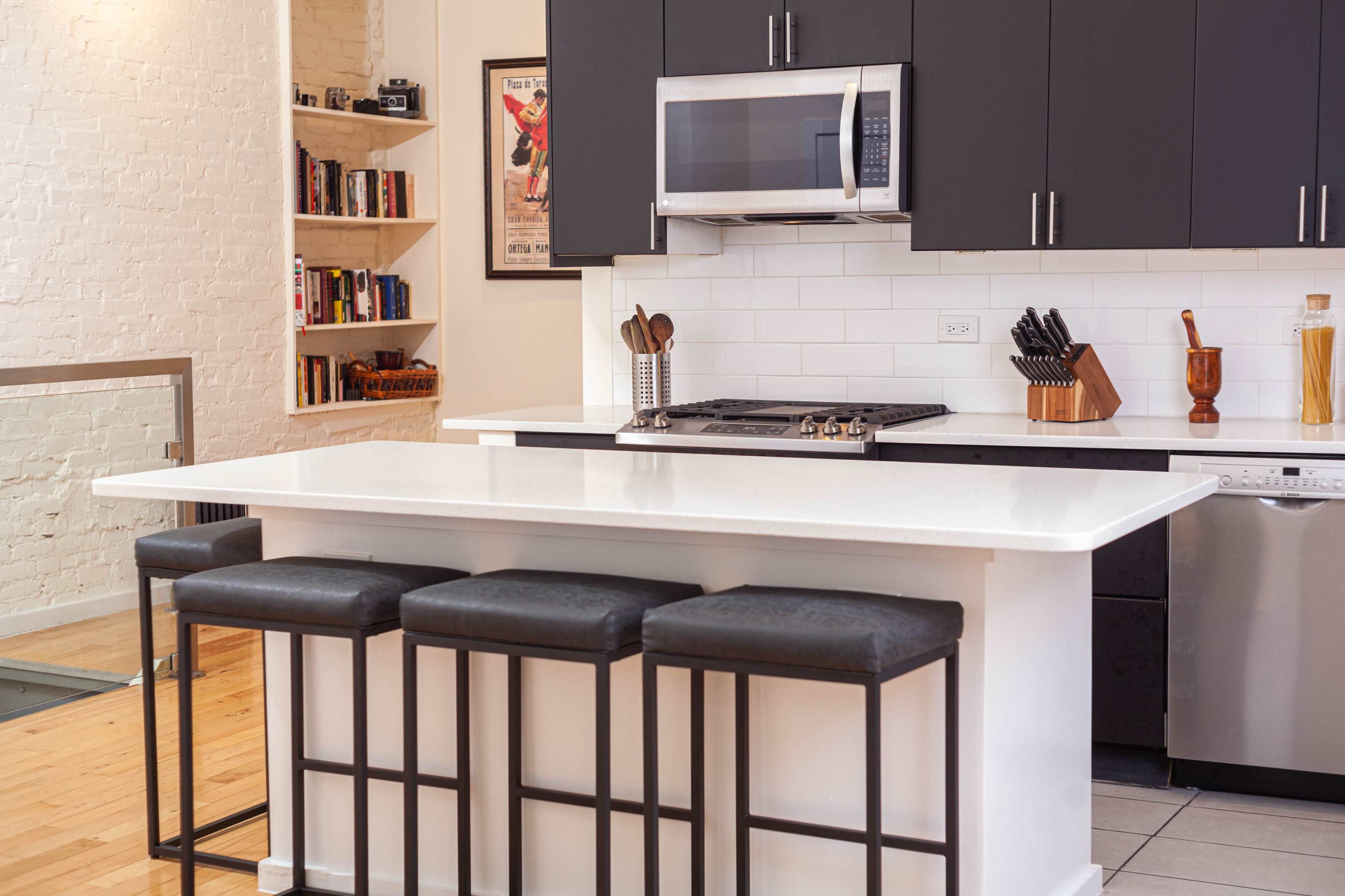 A modern kitchen featuring a white island with three black stools, stainless steel appliances, and dark cabinetry.