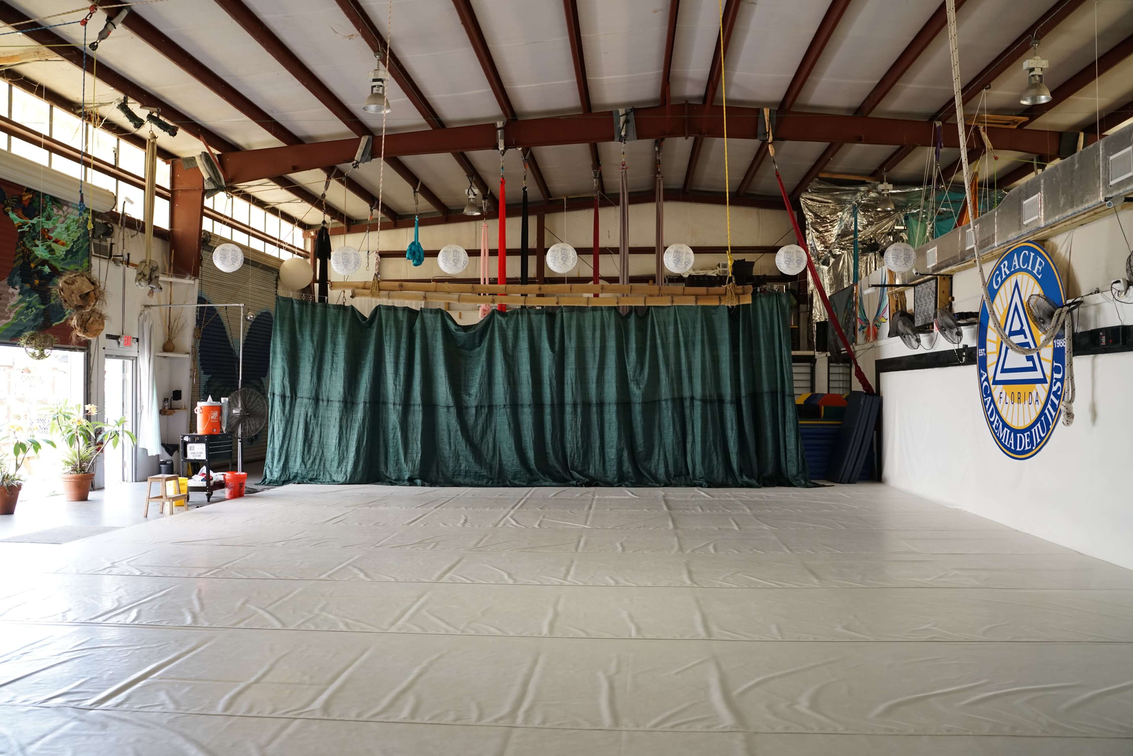 The image shows an empty indoor space with a large white mat on the floor, green curtains at the back, and a blue emblem on the wall.