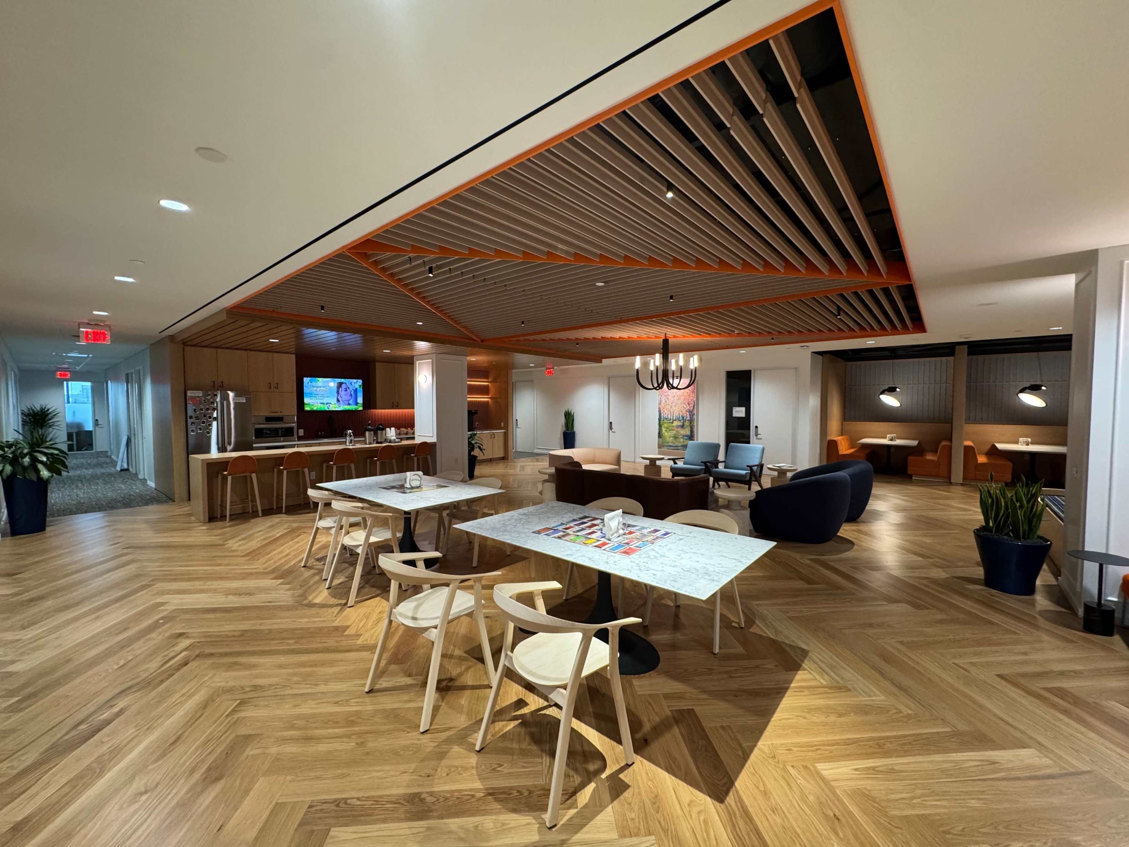 The image shows a modern office lobby area featuring a dining table with chairs, a kitchen space, and lounge seating under a decorative ceiling design with wooden slats.