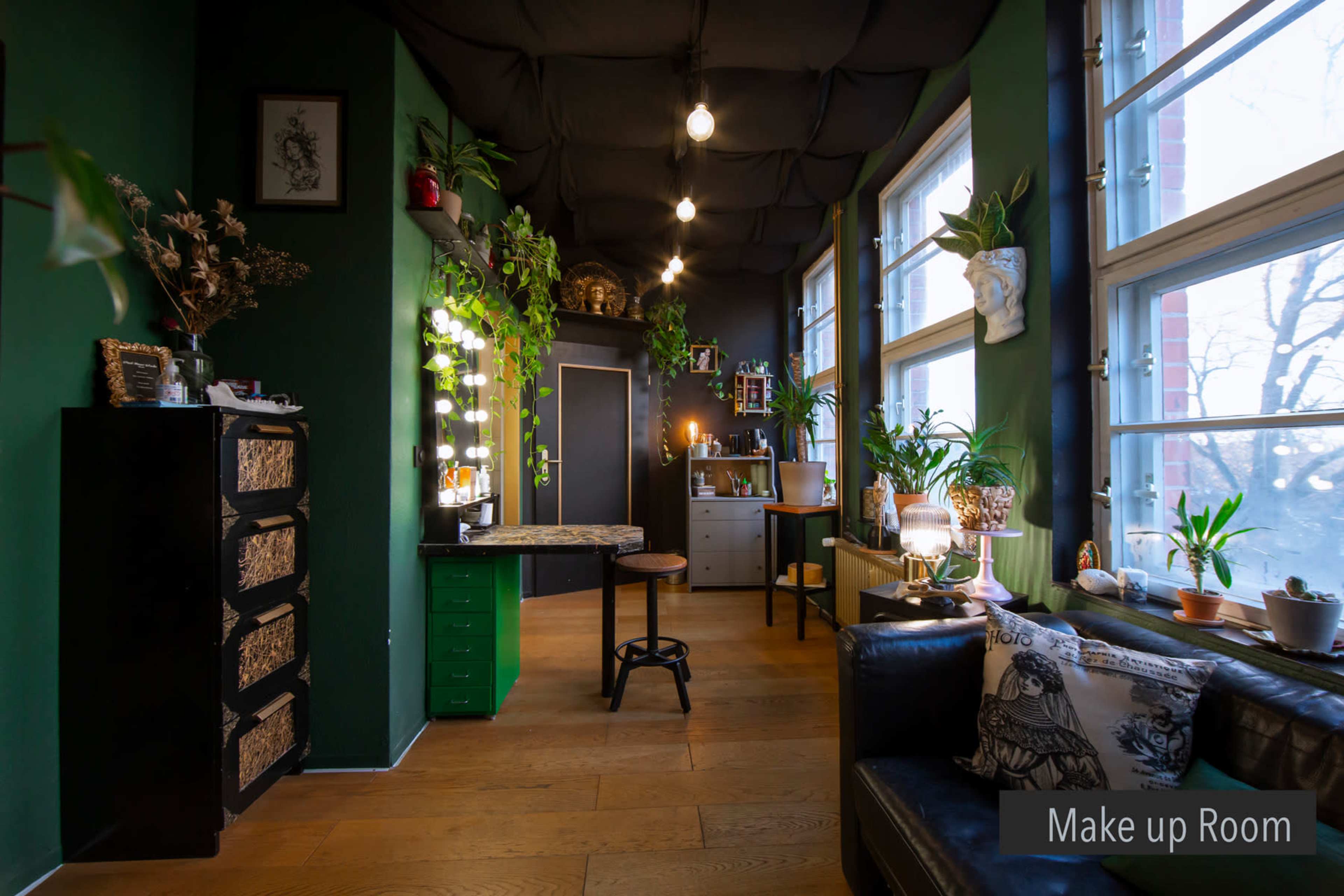 A makeup room featuring a dark green color scheme, a wooden floor, and multiple plants, with a large mirror framed by lights on one wall.