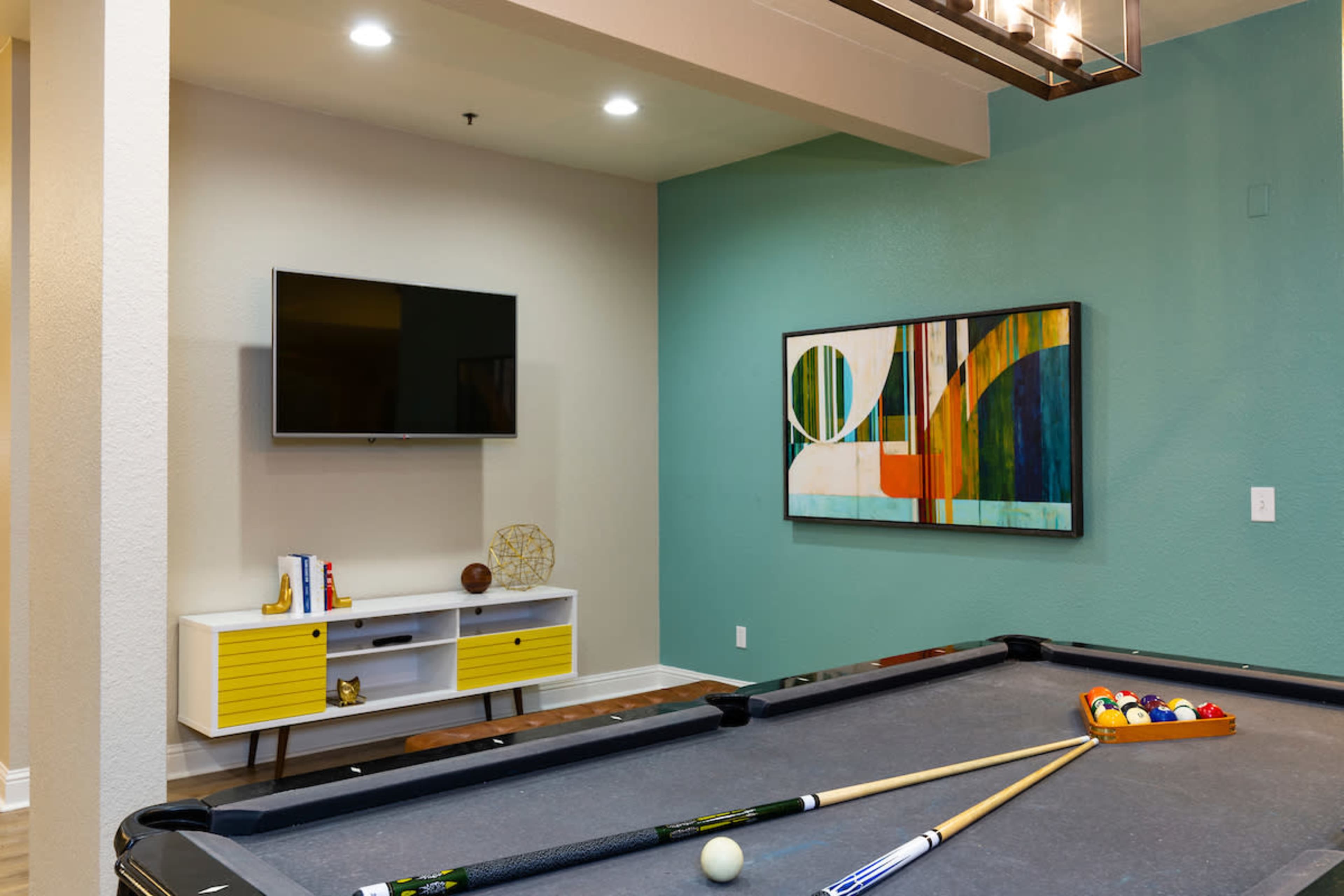 A pool table is positioned in a room featuring a television mounted on the wall and colorful artwork alongside a yellow and white media console.