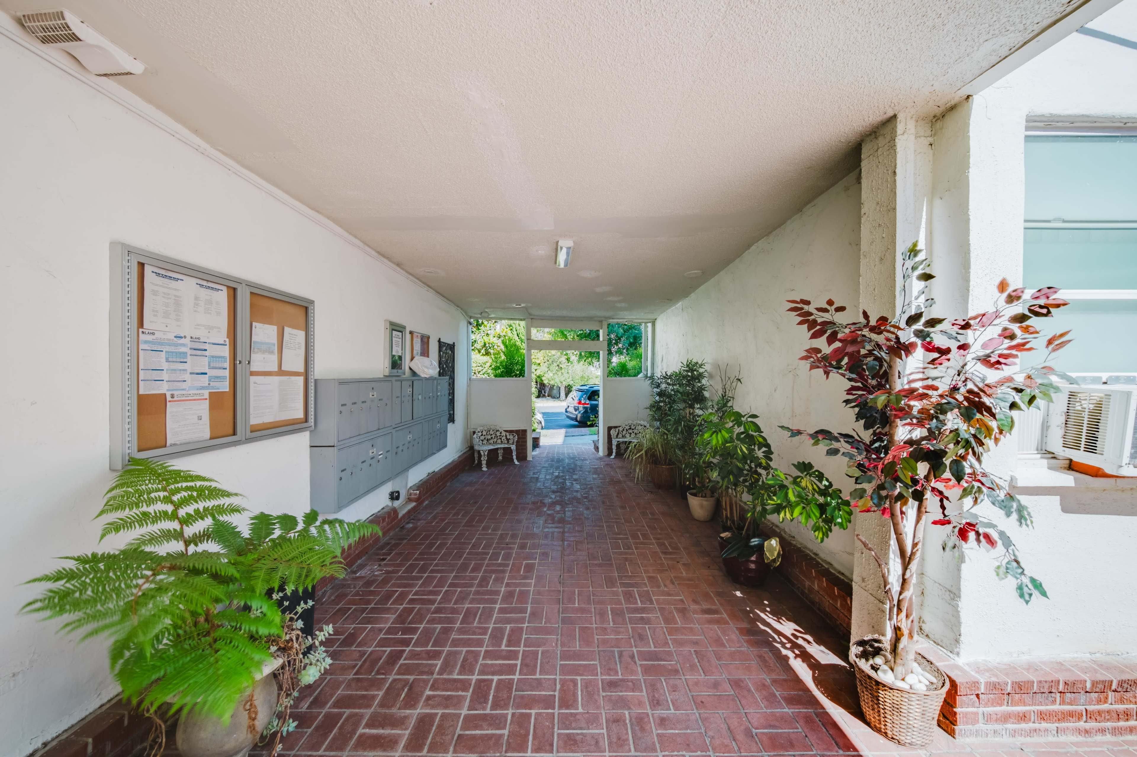 The image shows a tiled corridor with potted plants on either side and bulletin boards mounted on the wall.