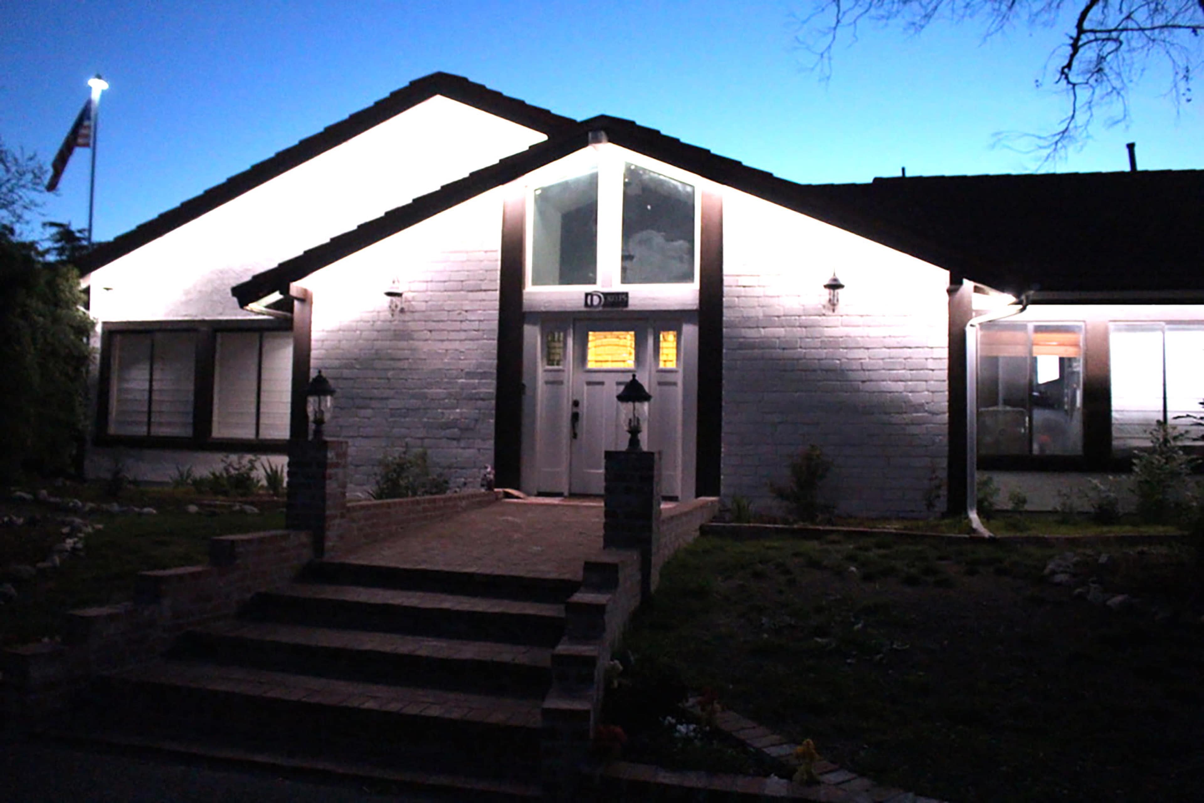 The image shows a well-lit house with a brick path leading to a front door, under a twilight sky.