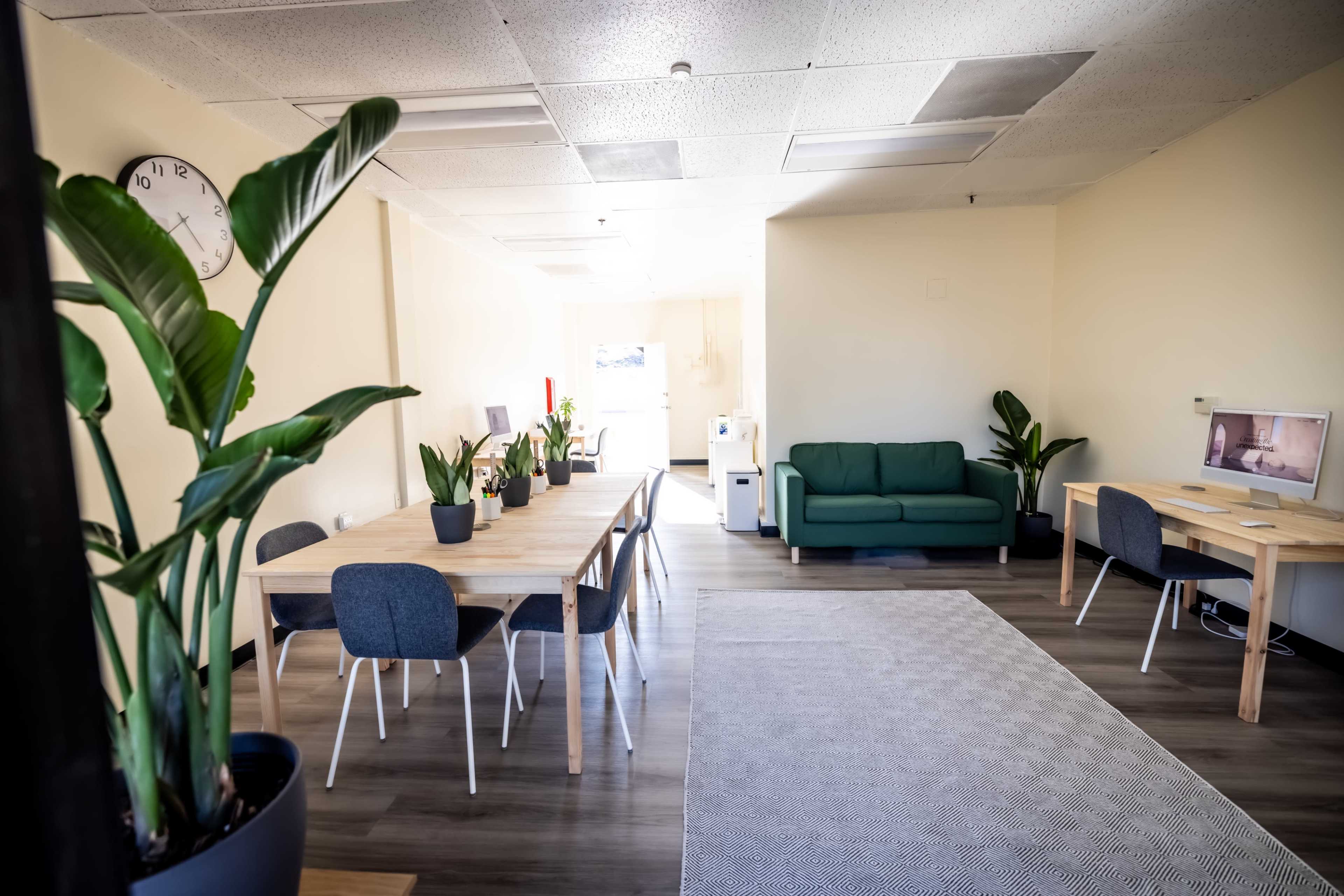 The image shows a bright office space featuring a green sofa, a dining table with chairs, and computer workstations, all adorned with potted plants.