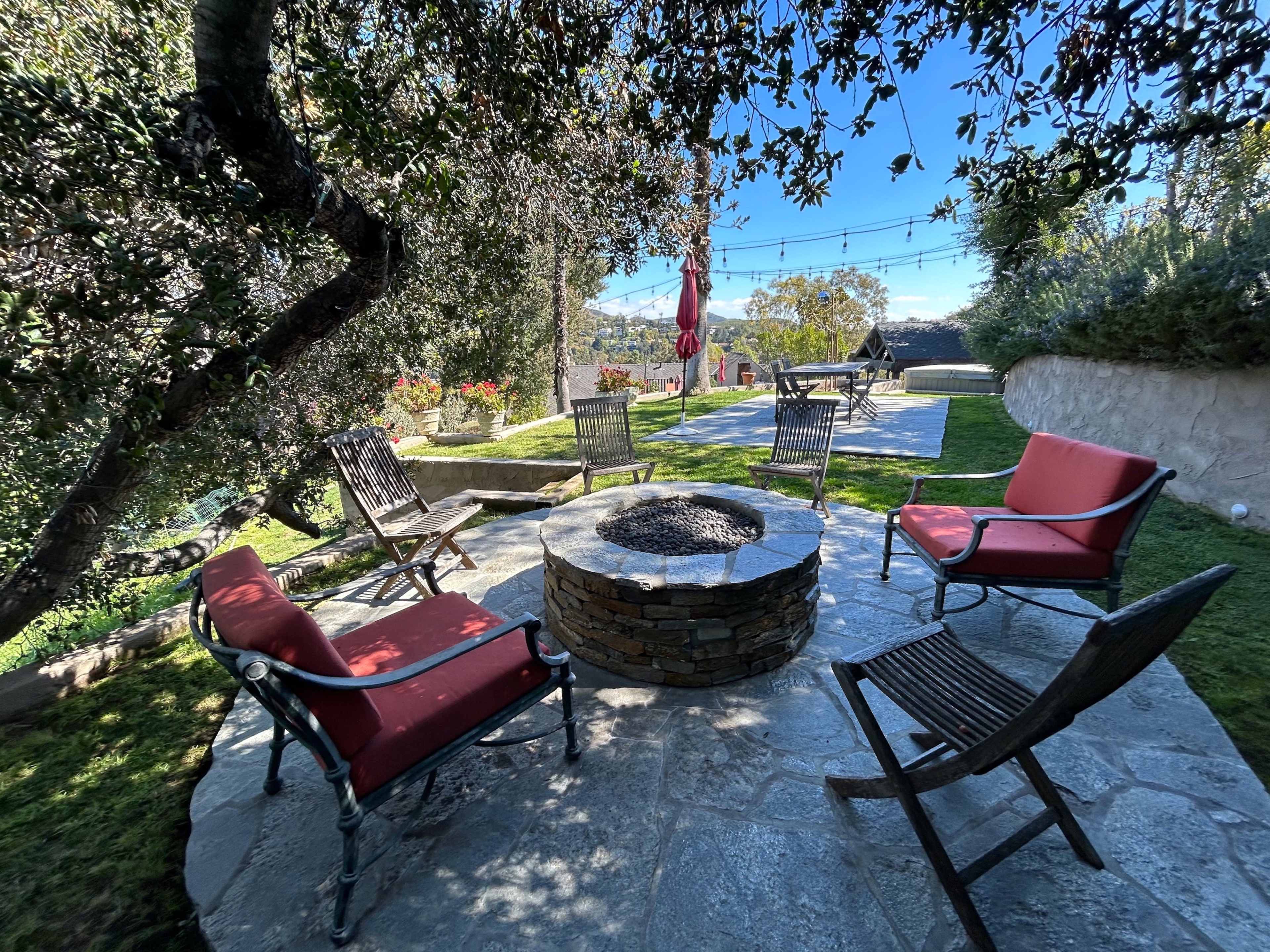 The image shows a circular stone fire pit surrounded by red and wooden chairs in a landscaped outdoor area with greenery and a clear blue sky.