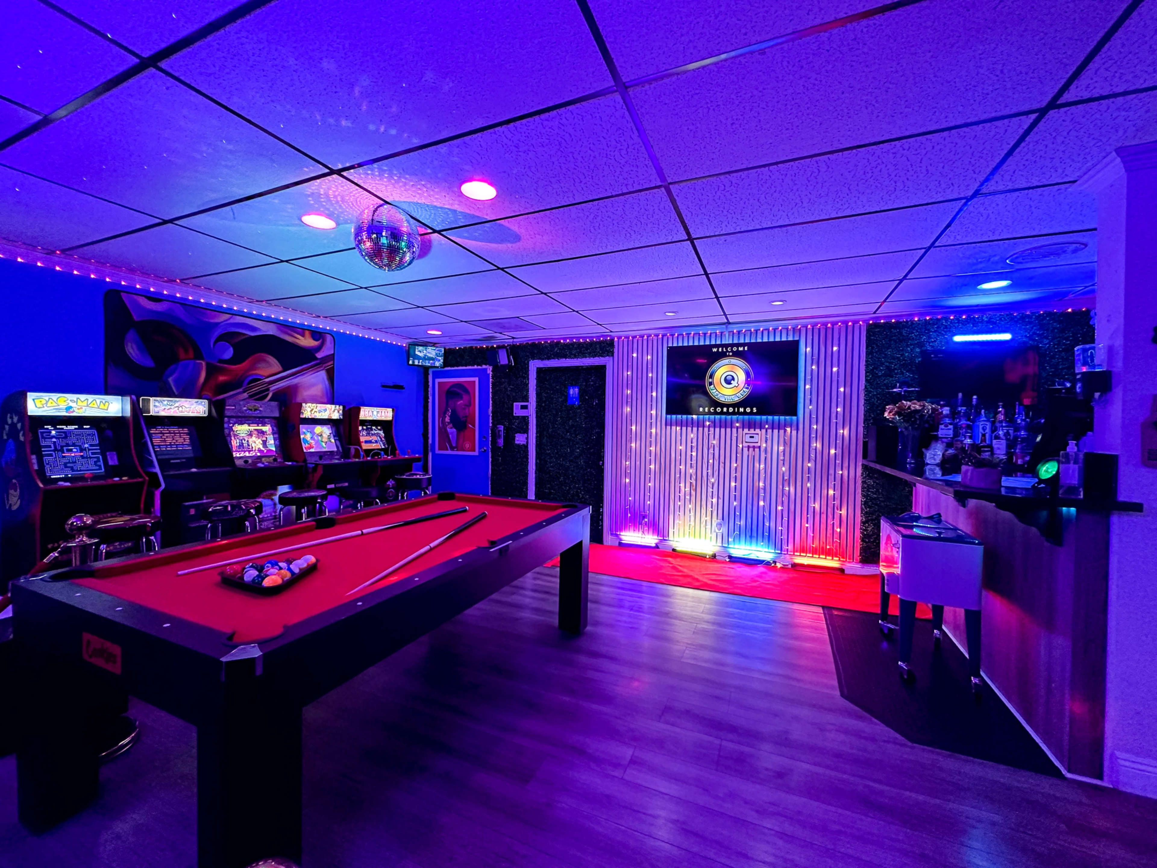 The image shows a vibrant game room featuring a pool table, arcade machines, a bar area, and colorful lighting.