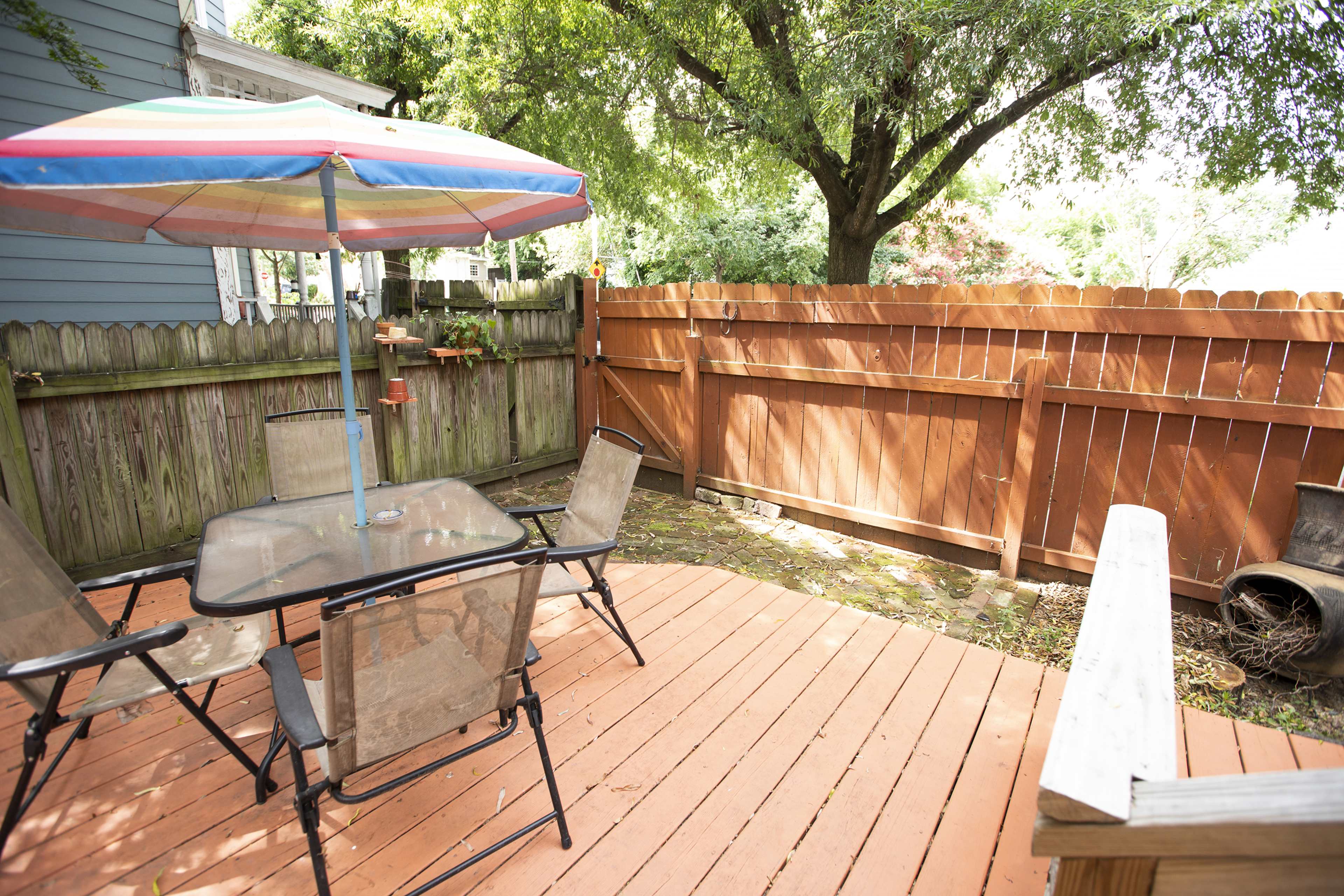 A wooden deck with a glass table and four chairs, shaded by a colorful umbrella, surrounded by a fenced yard and trees.