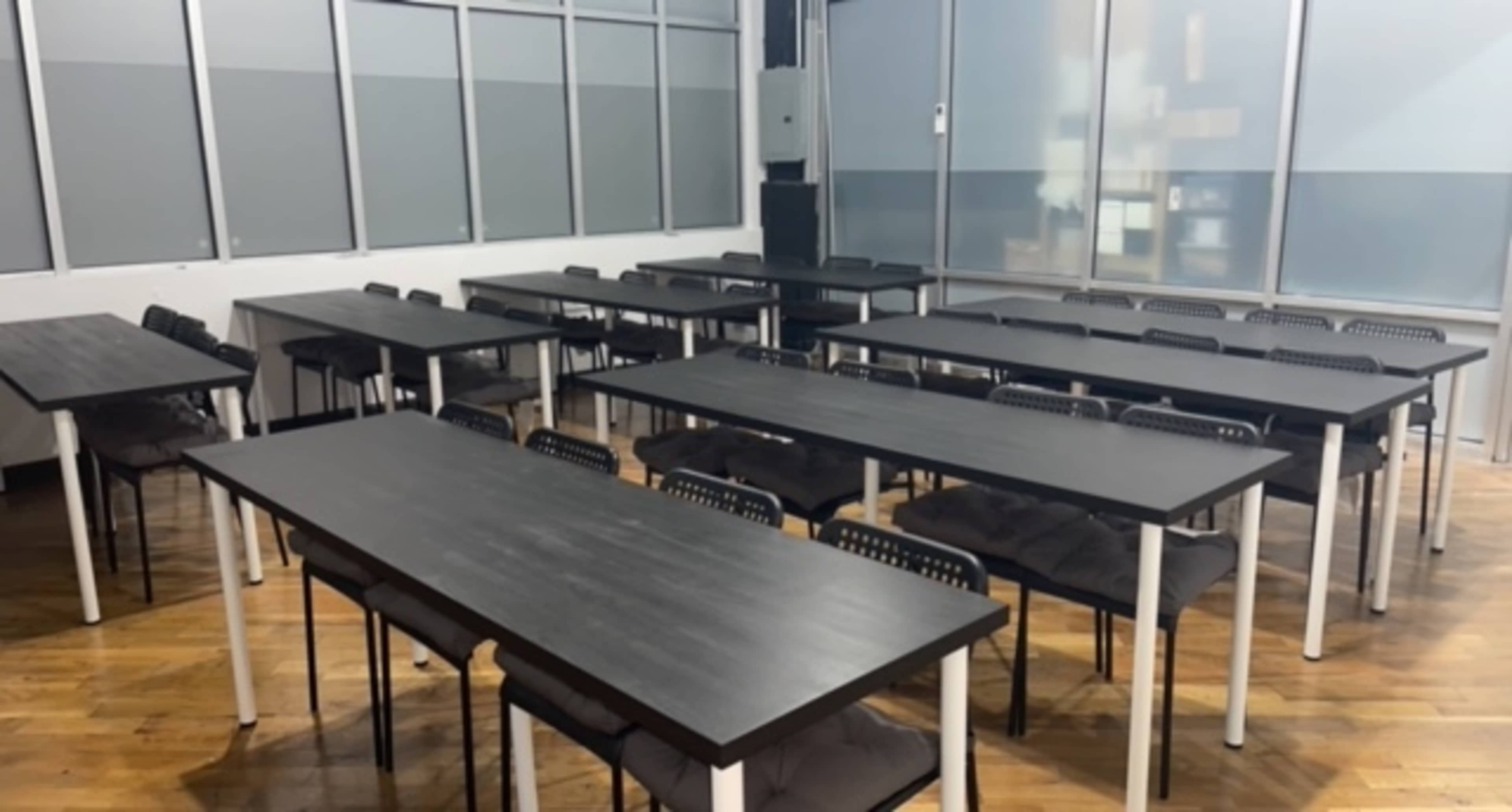 A classroom or meeting space arranged with multiple long tables and chairs, set up in rows on a wooden floor.