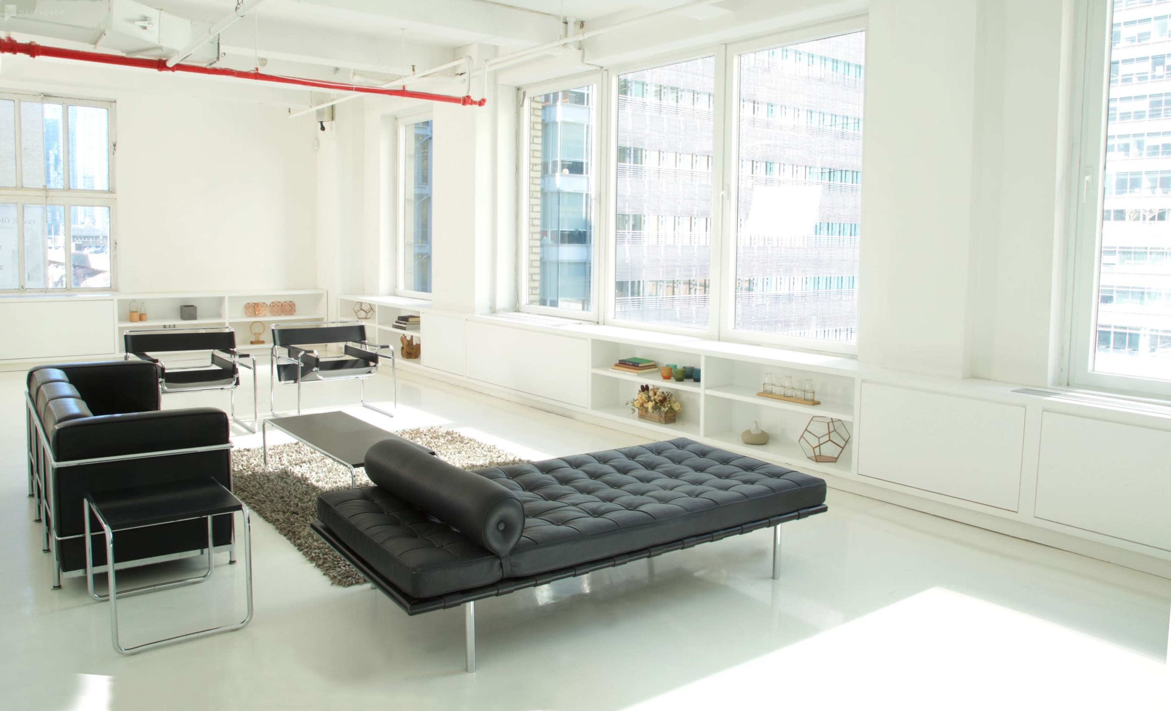 The image depicts a modern, minimalist office space with large windows, a black lounge chair, and a tufted daybed positioned on a rug.
