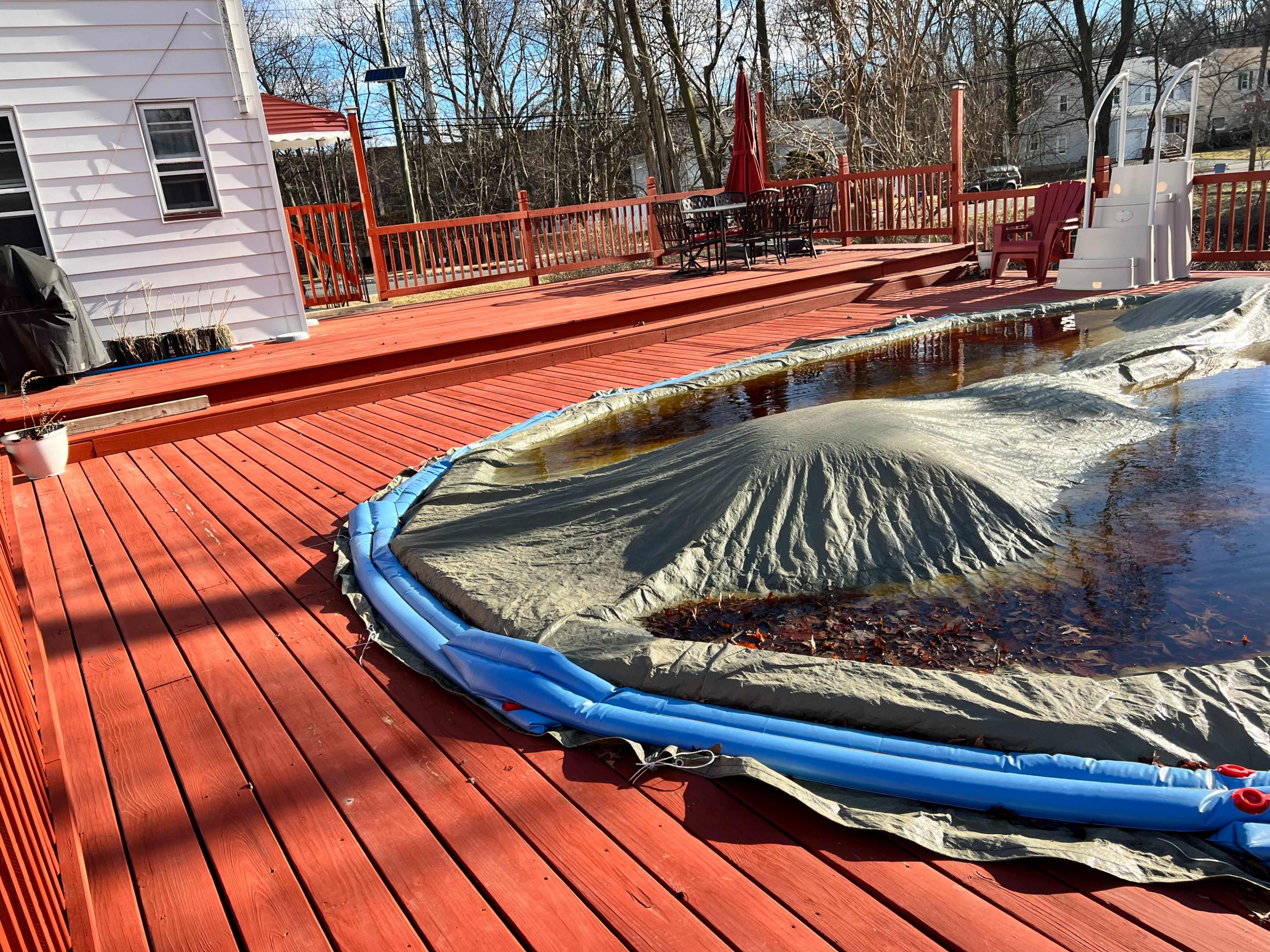 A covered swimming pool sits on a red wooden deck, surrounded by trees and patio furniture.