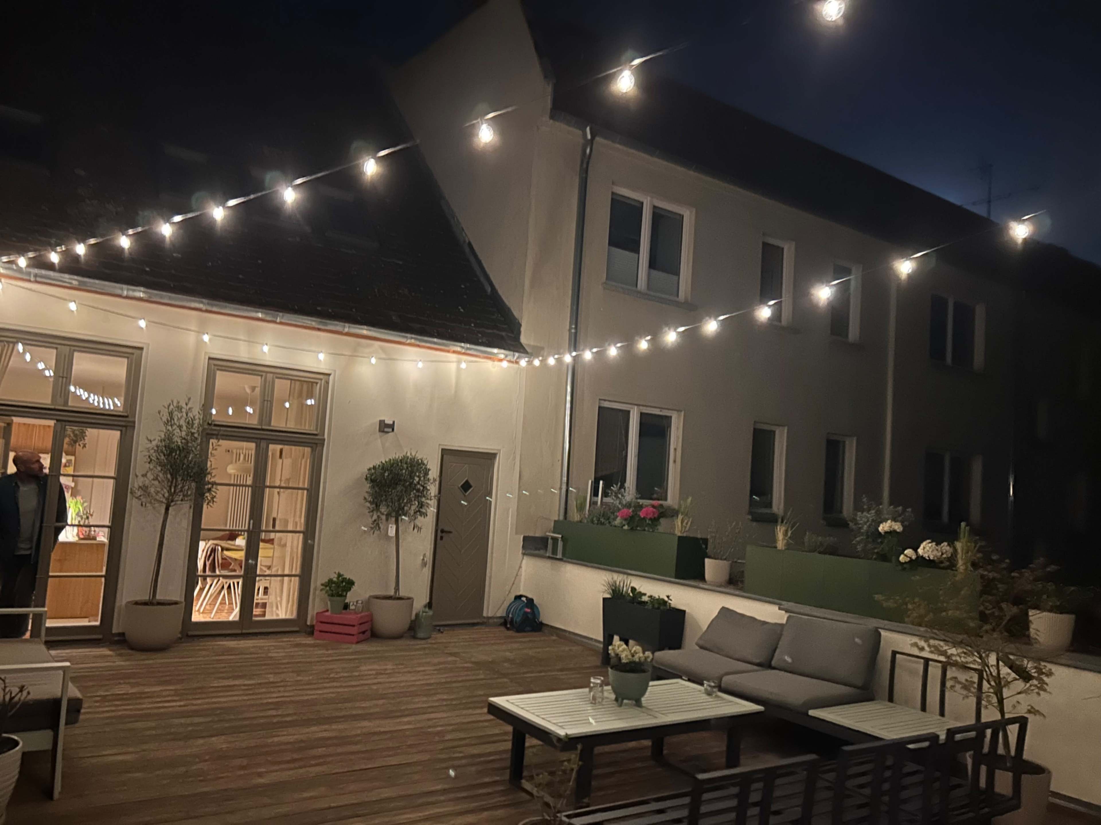 A well-lit patio area features string lights overhead, with potted plants and outdoor seating arranged against the backdrop of a two-story building.