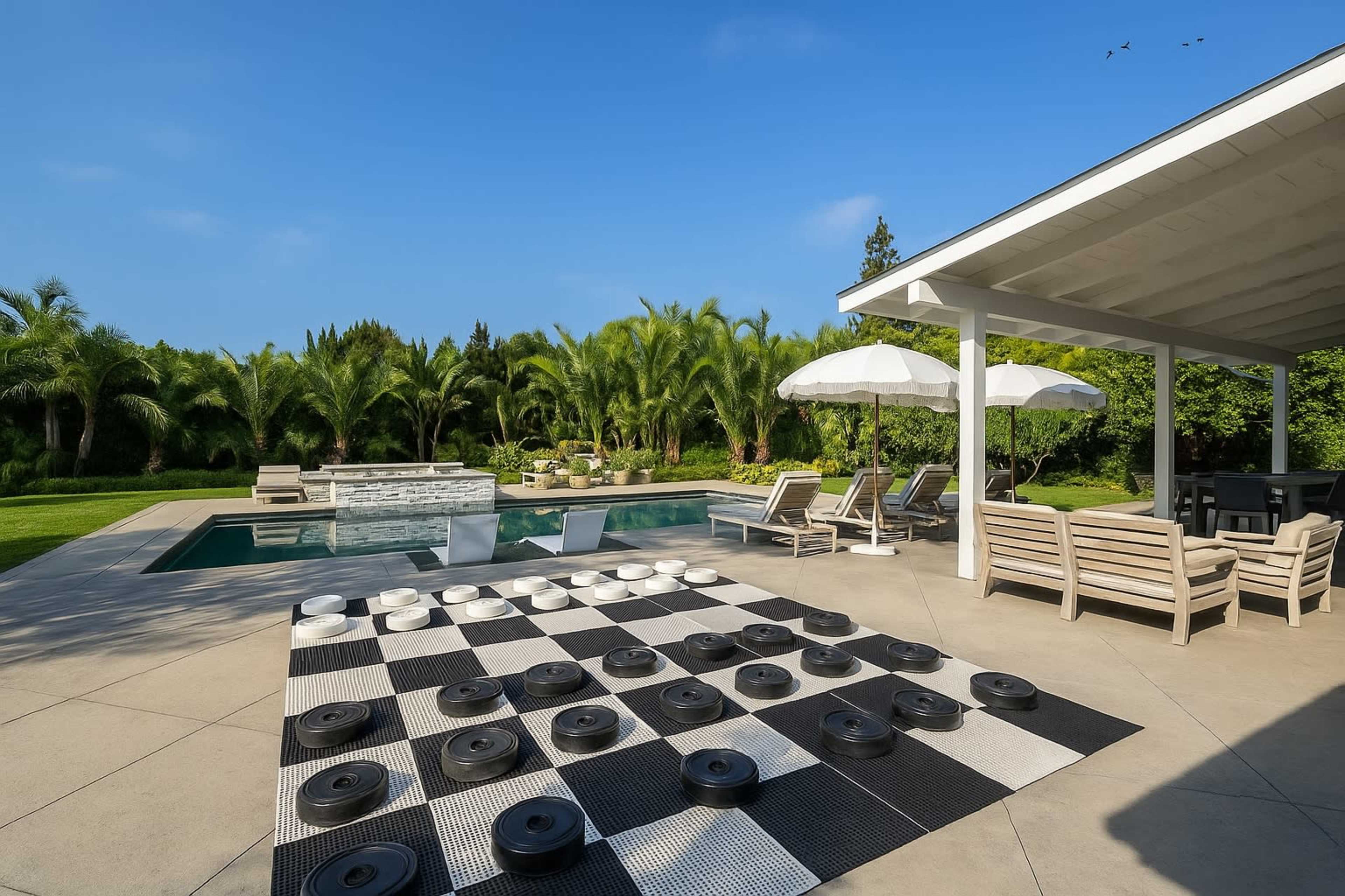 A checkers board is set up beside a swimming pool surrounded by lounge chairs and palm trees.