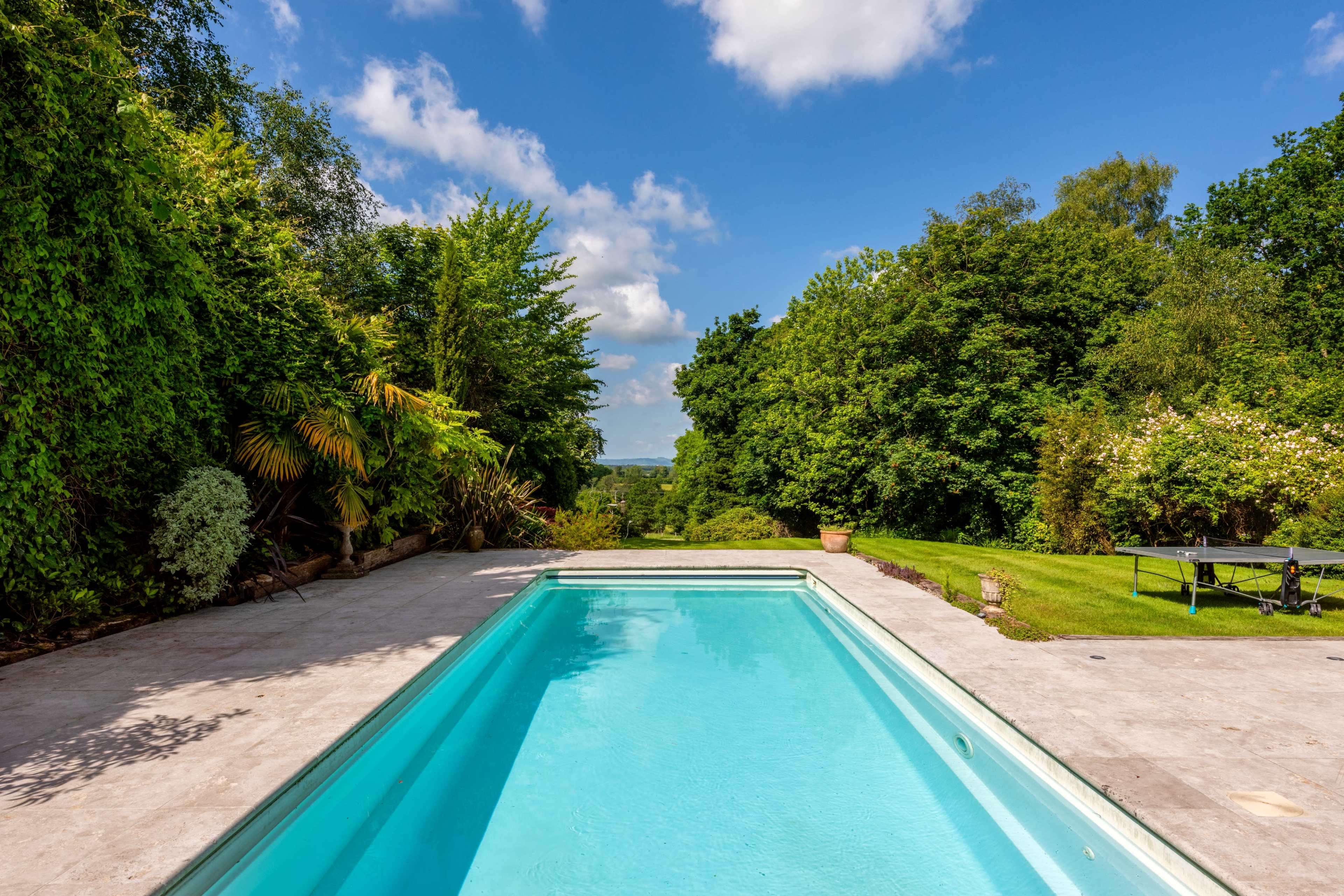 A clear swimming pool is surrounded by green foliage and grass under a partly cloudy sky.