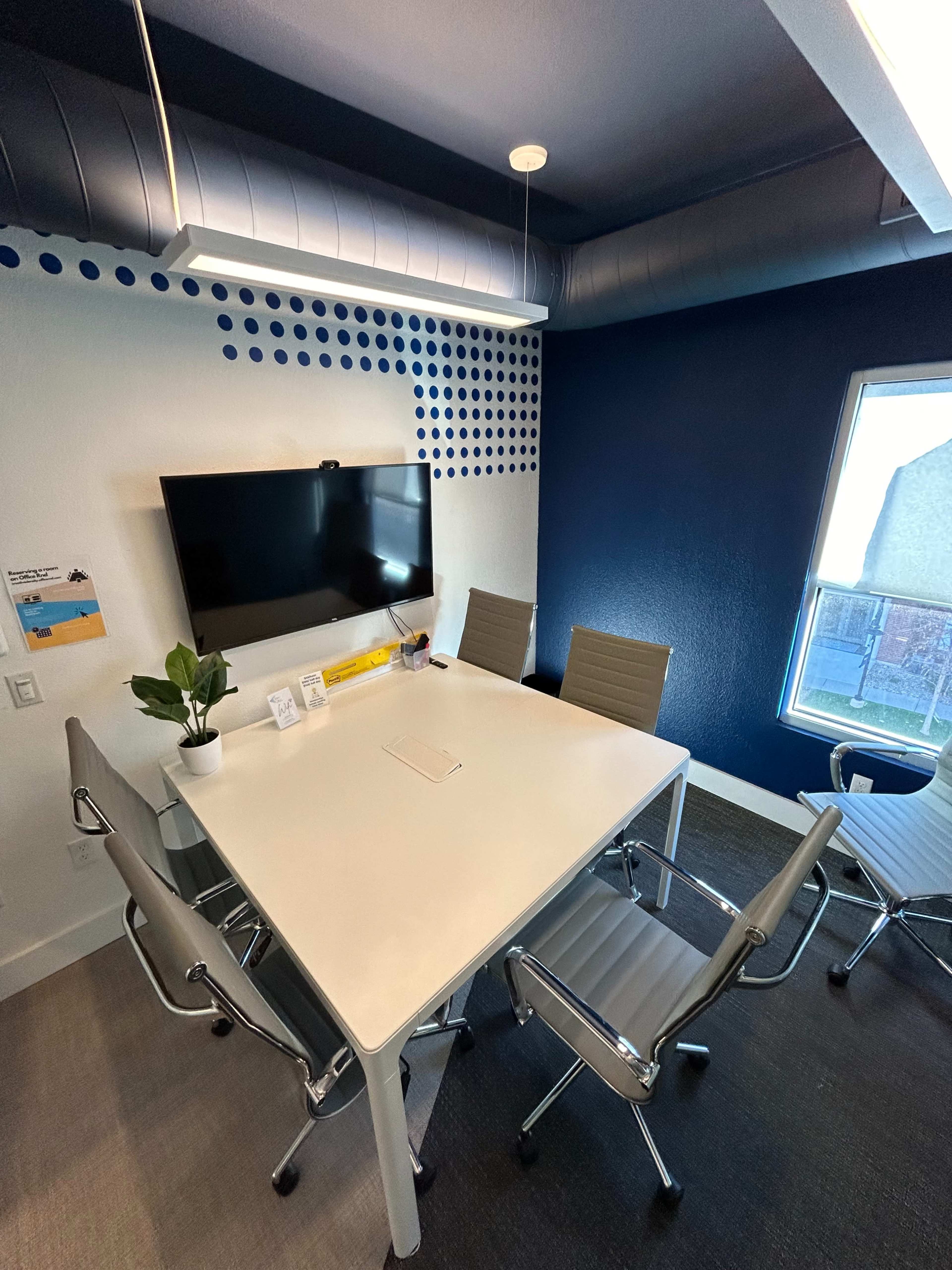 A small conference room with a white table, four metal chairs, a wall-mounted television, and a window.