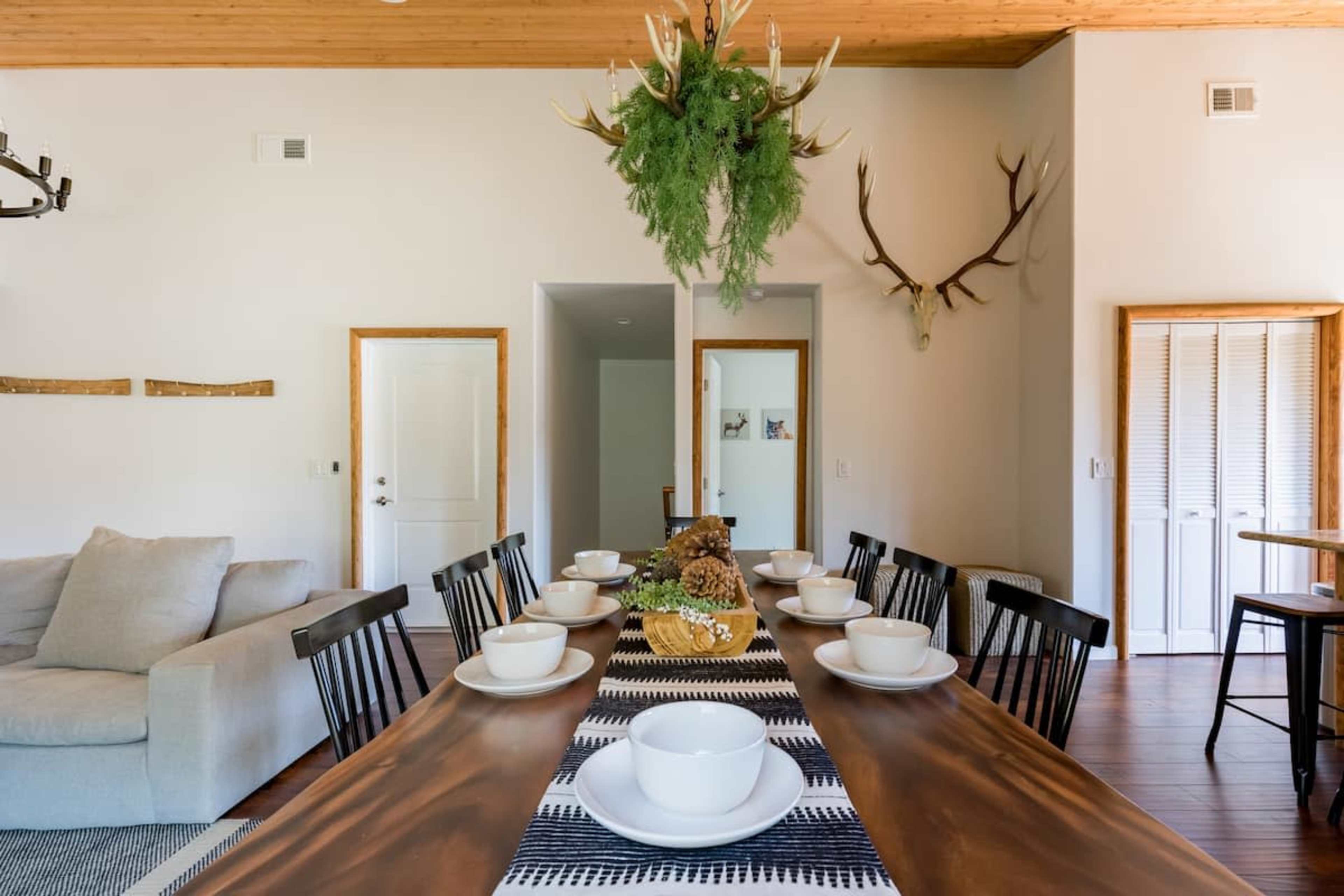 A long wooden dining table is set with white bowls and centered with a decorative arrangement, while a simple living area is visible in the background.